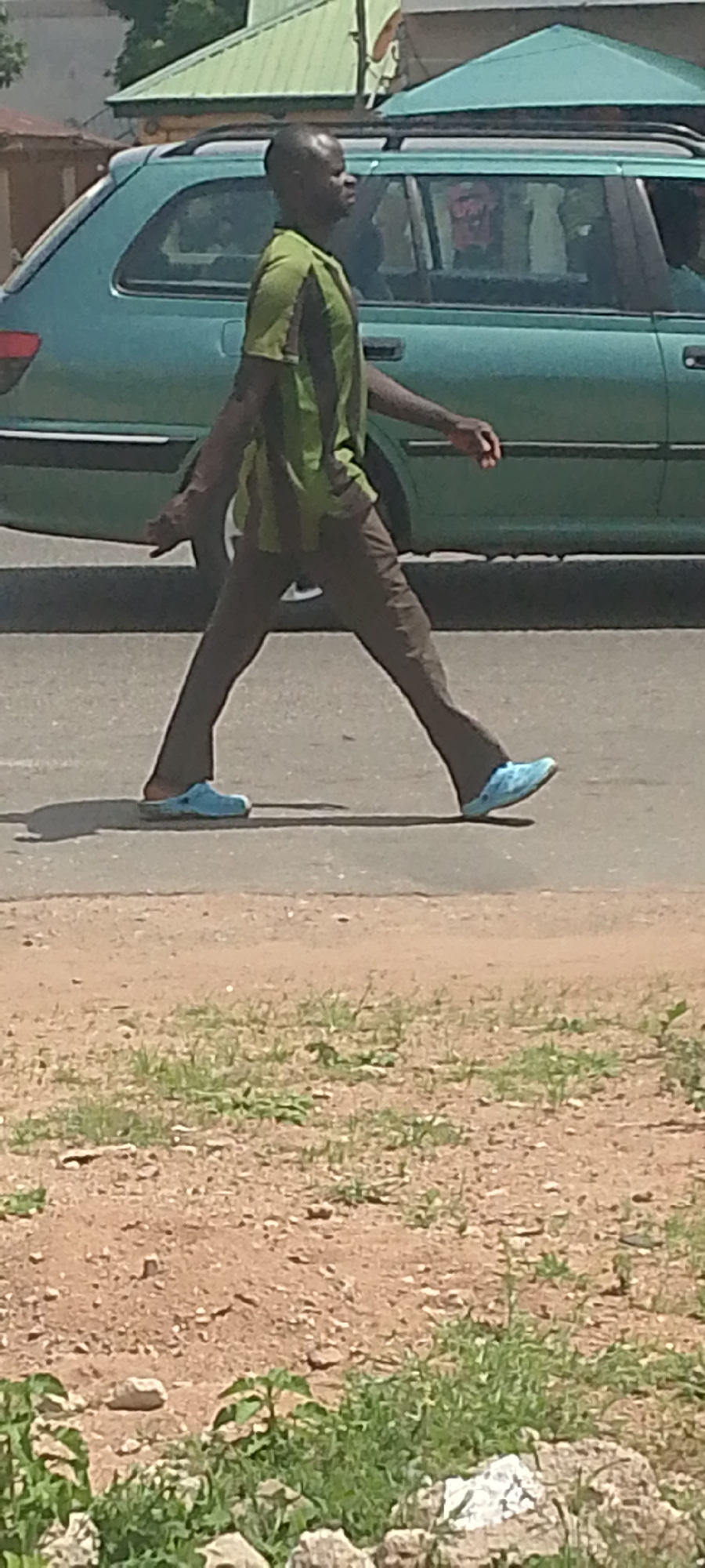 A man walks across a road in Jalingo, Nigeria. He is wearing a green and dark striped t-shirt and brown pants. He has light blue clogs on his feet. Behind him, a teal-colored station wagon is driving on the road, and there are some buildings and trees in the background. The scene appears to be outdoors during the daytime, with the sun casting shadows on the ground. The weather seems to be clear, as indicated by the bright sunlight. The man is walking with a determined stride, and his expression is neutral. The setting appears to be an urban or semi-urban environment, given the presence of the road, car, and buildings. There is no visible text in the image.