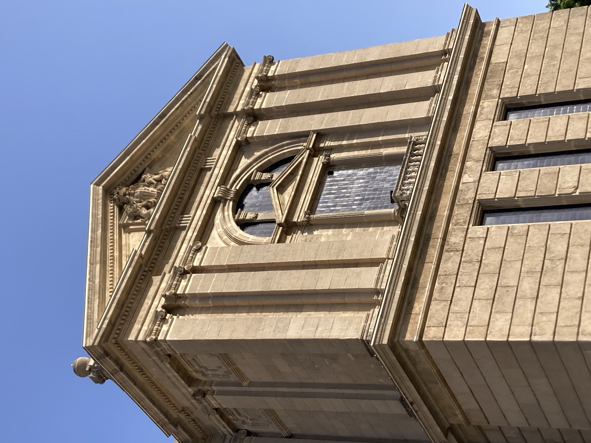 The image shows a close-up, upward-facing view of the facade of the Palau de la Generalitat de Catalunya in Barcelona, Spain, under a clear blue sky. The building is constructed of light-colored stone, with classical architectural elements such as pilasters, cornices, and decorative carvings. The facade is adorned with multiple windows, including a large arched window with detailed stonework above it. Below the arched window, there is a small balcony with a railing. The building displays a symmetrical design with a prominent triangular pediment at the top, which features a coat of arms. The stone blocks forming the lower portion of the facade are clearly visible, laid in horizontal courses. The overall impression is one of historical grandeur and architectural solidity.