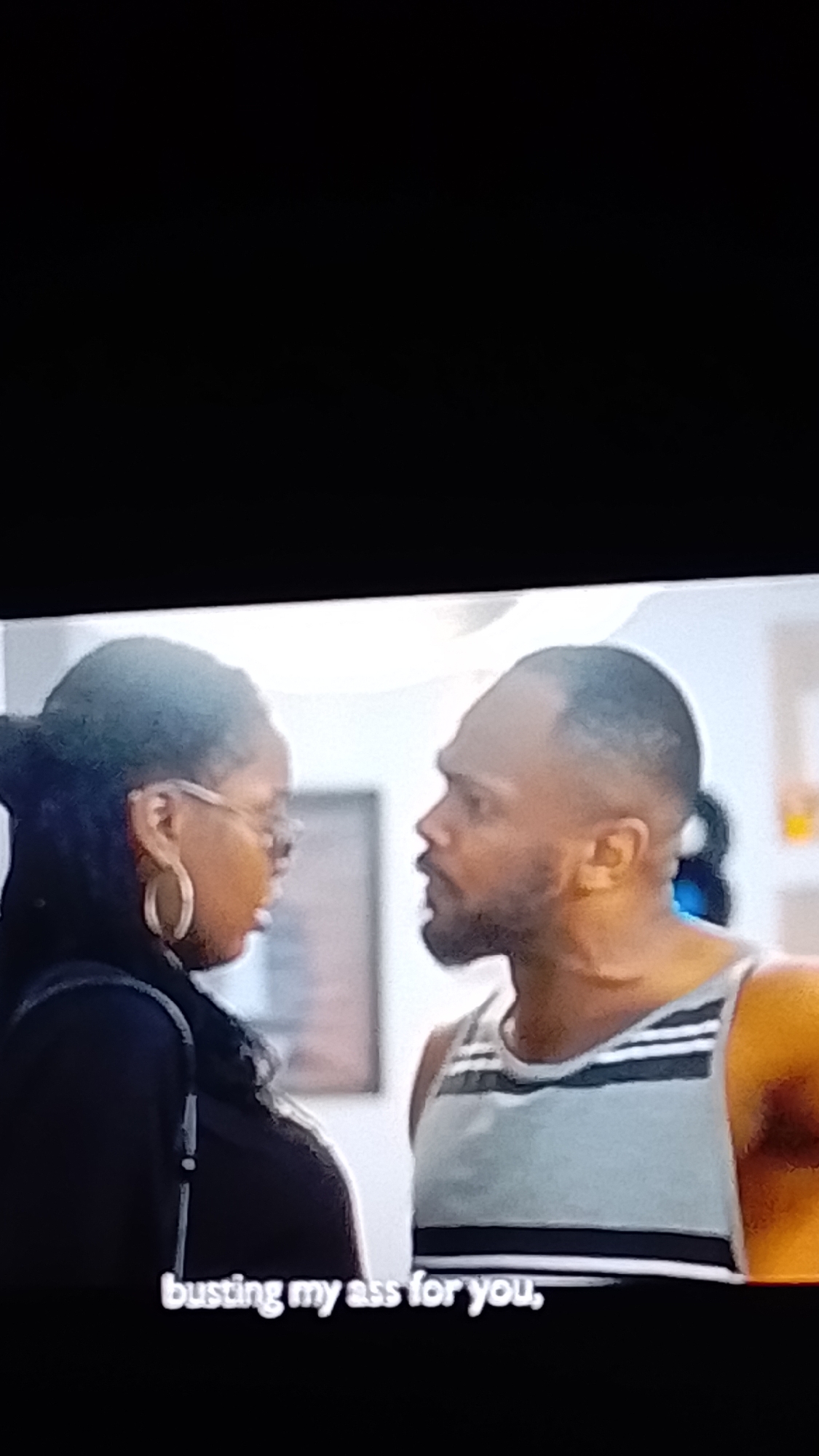 The image shows a close-up of two people, a woman and a man, facing each other. The woman, on the left, has dark curly hair and is wearing large hoop earrings and glasses. She is looking towards the man with a serious expression. The man, on the right, has short hair and a beard, and is wearing a grey tank top with horizontal black and white stripes. He appears to be speaking or yelling, with his mouth open. The background is blurred but suggests an indoor setting with some objects that are not clearly identifiable. The lighting is bright, and there are no visible weather cues. The overall mood appears to be one of tension or argument.
At the bottom of the image, there is visible text that reads: "busting my ass for you,". This text suggests a dialogue or narration related to the scene. The location context provided is Lokoja, Nigeria, but there are no specific visual cues in the image to confirm this location.