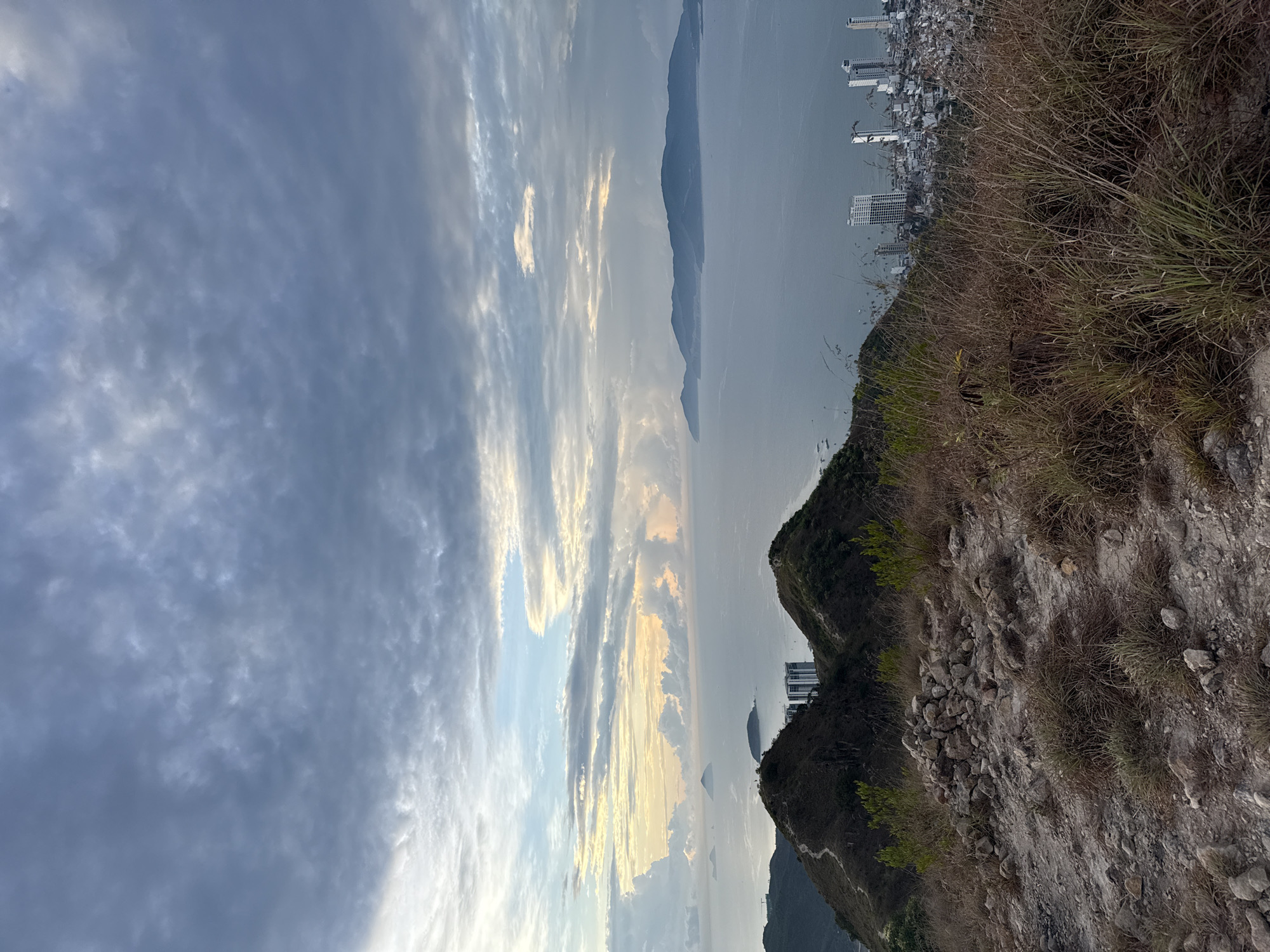 The image presents a landscape view from a high vantage point, possibly a mountain or cliff edge. The sky occupies a significant portion of the image, displaying a mix of clouds, some of which are illuminated with a golden hue, suggesting sunrise or sunset. Below the sky, there is a body of water, possibly a bay or ocean, with a mountain range visible in the distance. A cityscape with tall buildings is visible on the shoreline, indicating a populated area. The foreground features rocky terrain with sparse vegetation, typical of a hillside or cliffside environment. Given the location provided (Bac Nha Trang Ward, Vietnam), the cityscape is likely Nha Trang.