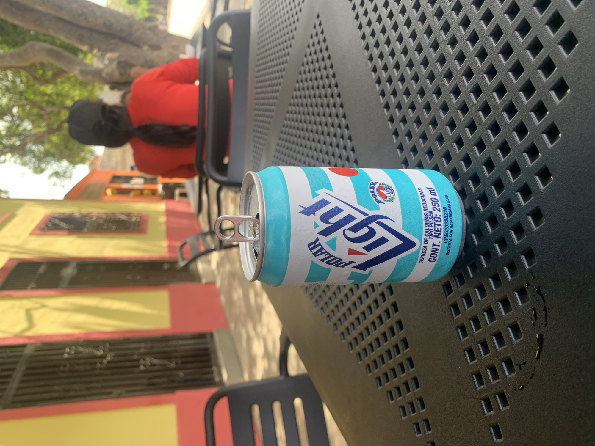 The image shows an open can of Polar Light beer on a black metal table with a lattice pattern. In the background, partially obscured by the table, a person wearing a red shirt and black cap sits in a chair. Behind the person, a yellow and pink building is visible. The angle of the shot indicates the photographer is seated at the same table. Based on the writing in Spanish on the can, the location is likely Santa Ana, Venezuela.