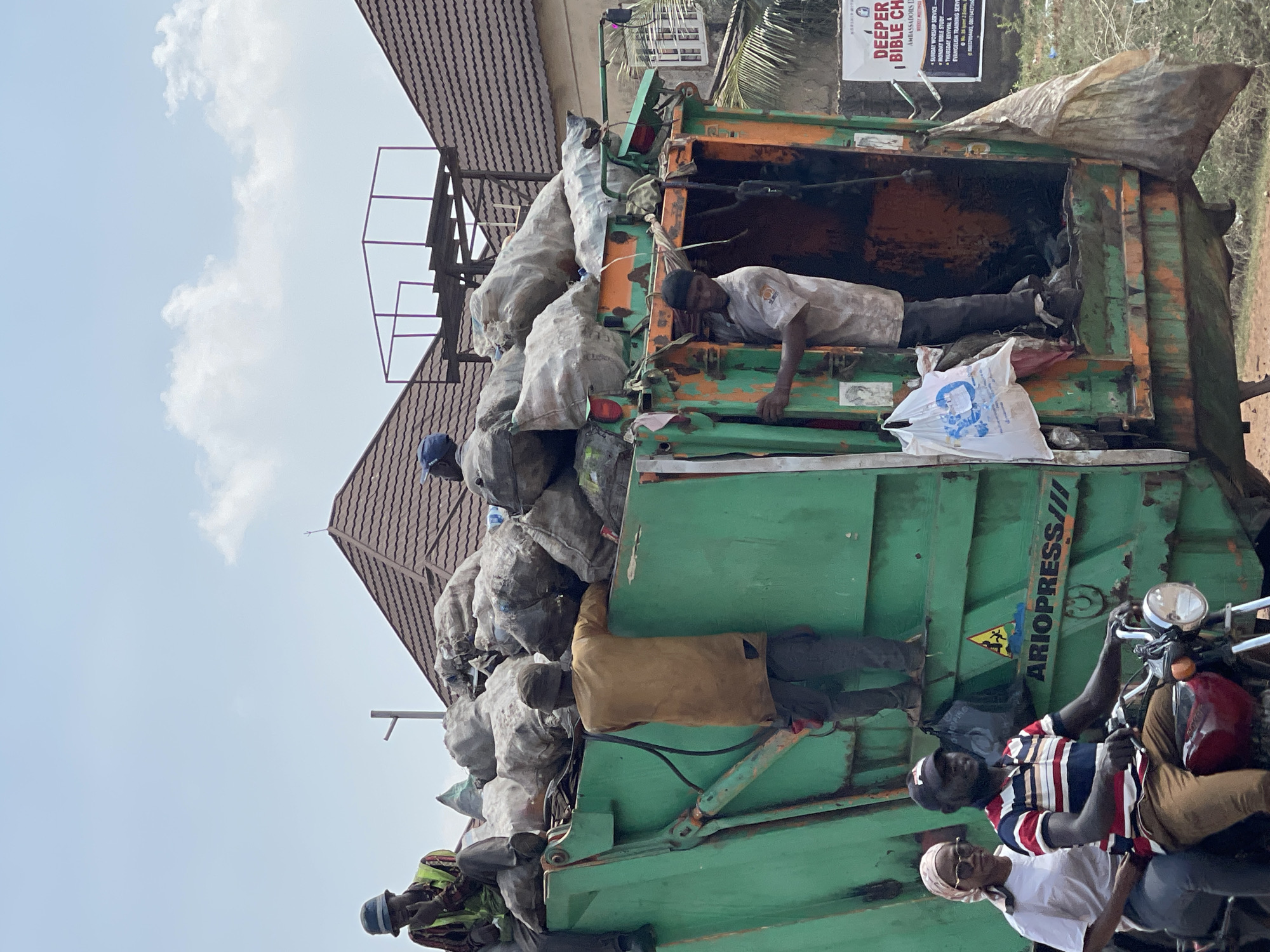 The image shows a heavily loaded green and orange garbage truck on a street in Abuja, Nigeria. The truck bed is filled with bulging bags. Three individuals are riding on the outside of the truck on the green area. One person is inside the orange trash bin. A man and a woman are sitting on a motorcycle near the truck. A building is visible in the background, and a sign is placed on a pole. The text on the sign is “DEEPER BIBLE CH”. The environment appears to be a mixed residential and commercial area on a sunny day.