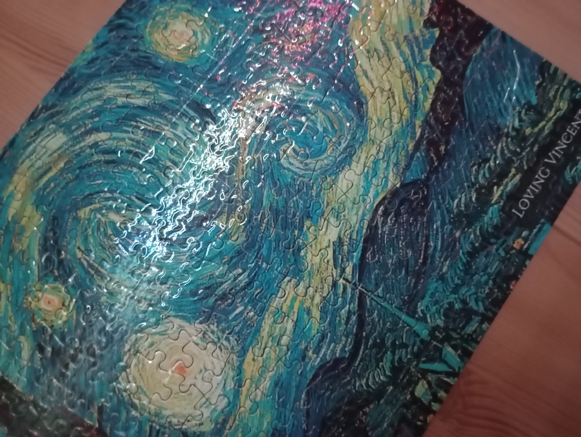 The image shows a jigsaw puzzle partially completed, depicting Vincent van Gogh's "The Starry Night" painting. The puzzle pieces form swirling patterns of blue, yellow, and white colors, representing the night sky and stars. On the right side of the puzzle, near the bottom, the text "LOVING VINCENT" is visible. The puzzle is placed on a wooden surface.