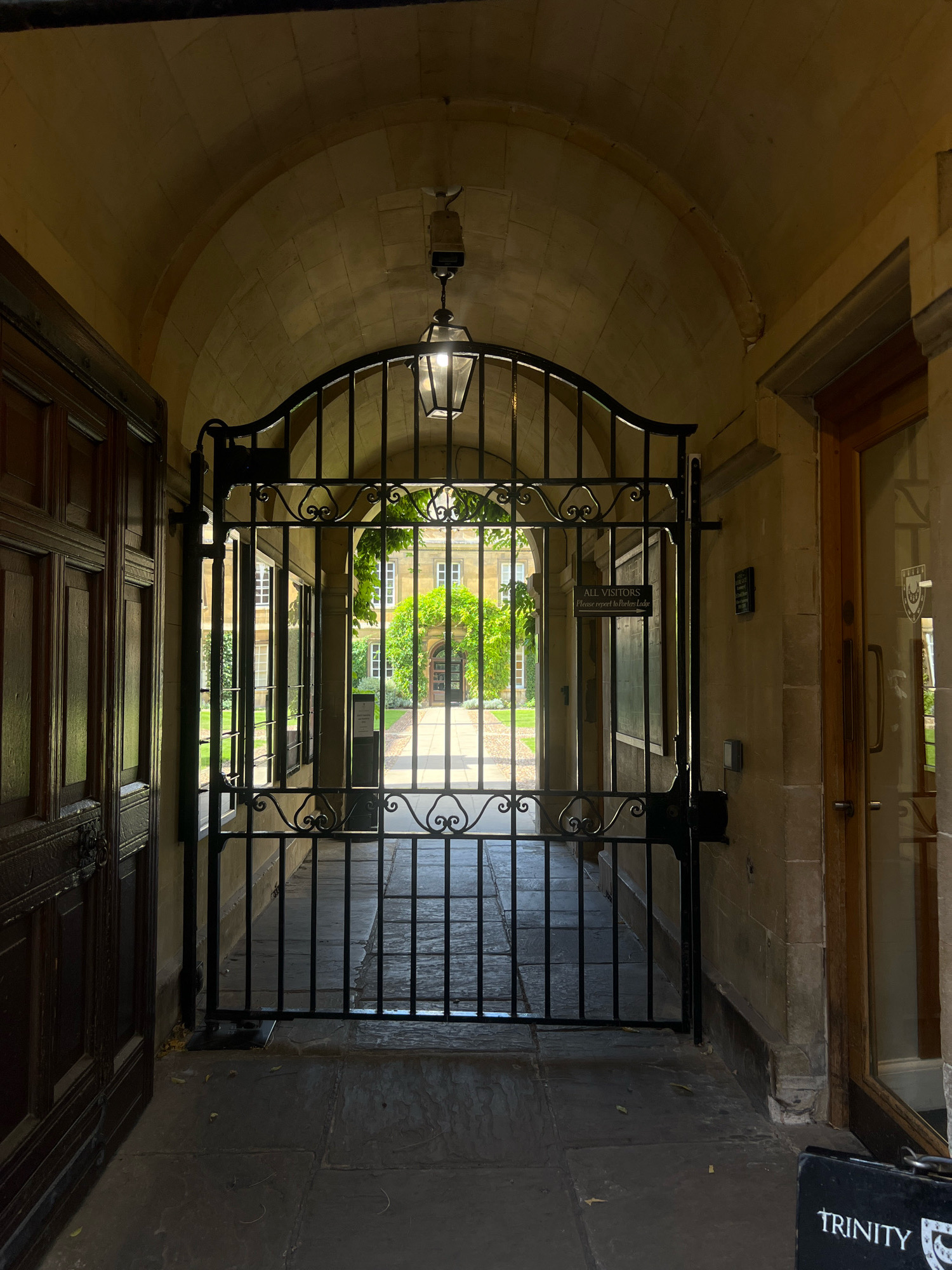The image shows a wrought-iron gate in an arched stone passageway, looking out onto a courtyard with a building in the distance. The gate is black and ornate, with scrollwork at the top and bottom. A lantern hangs from the ceiling of the archway. To the left of the gate is a large wooden door with carved panels. To the right is a glass door with a wooden frame, and a sign on the wall above it. A smaller sign is attached to the gate itself. The courtyard is paved and has grass on either side. The building in the distance has multiple windows. On the pavement in the foreground, a black sign with "TRINITY" written on it is visible. This is an exterior view of Trinity College, Cambridge, United Kingdom.