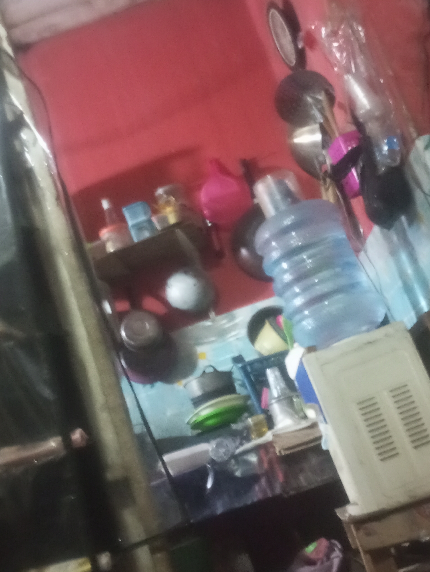 The image depicts a cluttered shelf or tabletop within an indoor environment. The background is a solid red wall. On the shelf, several items are visible, including what appears to be a large blue water jug, a white appliance with a slatted front, various bottles and containers, and what looks like a pink spherical object. The scene is poorly lit and the image is out of focus, making precise identification of all objects challenging. There are no individuals or discernible actions taking place. The location is described as Cilegon, Indonesia.
