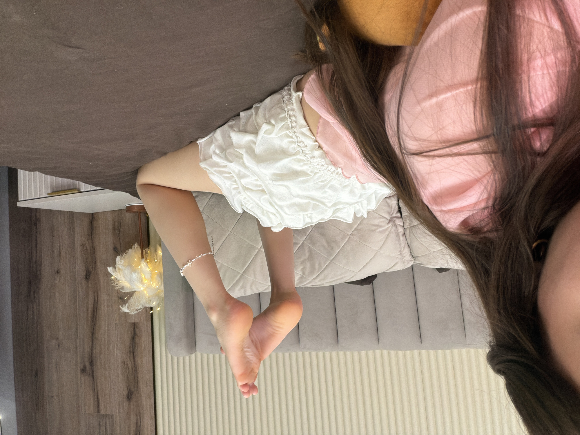 An individual is depicted lying face down on a bed in an indoor setting. The left side of the person's face, including a cheek and ear with a gold hoop earring, is visible in the immediate foreground, obscured by long, dark brown hair. The individual wears a pink top beneath their hair and white, ruffled shorts or underwear. Their bare legs are bent at the knees, with the soles of their feet raised upwards. A white, possibly pearl or beaded, ankle bracelet with a small floral charm adorns the right ankle. The bed is covered with a plain brown duvet or blanket. Light grey, quilted pillows are arranged on the bed. A grey upholstered headboard with vertical panels is visible behind the bed. To the right, a white bedside table with a ridged front and a gold-colored handle supports a decorative lamp featuring a white, feathery or floral shade with warm yellow illumination. The wall behind the headboard is light-colored with vertical striations, while a section of the wall to the right features a dark wood paneling or wallpaper.
