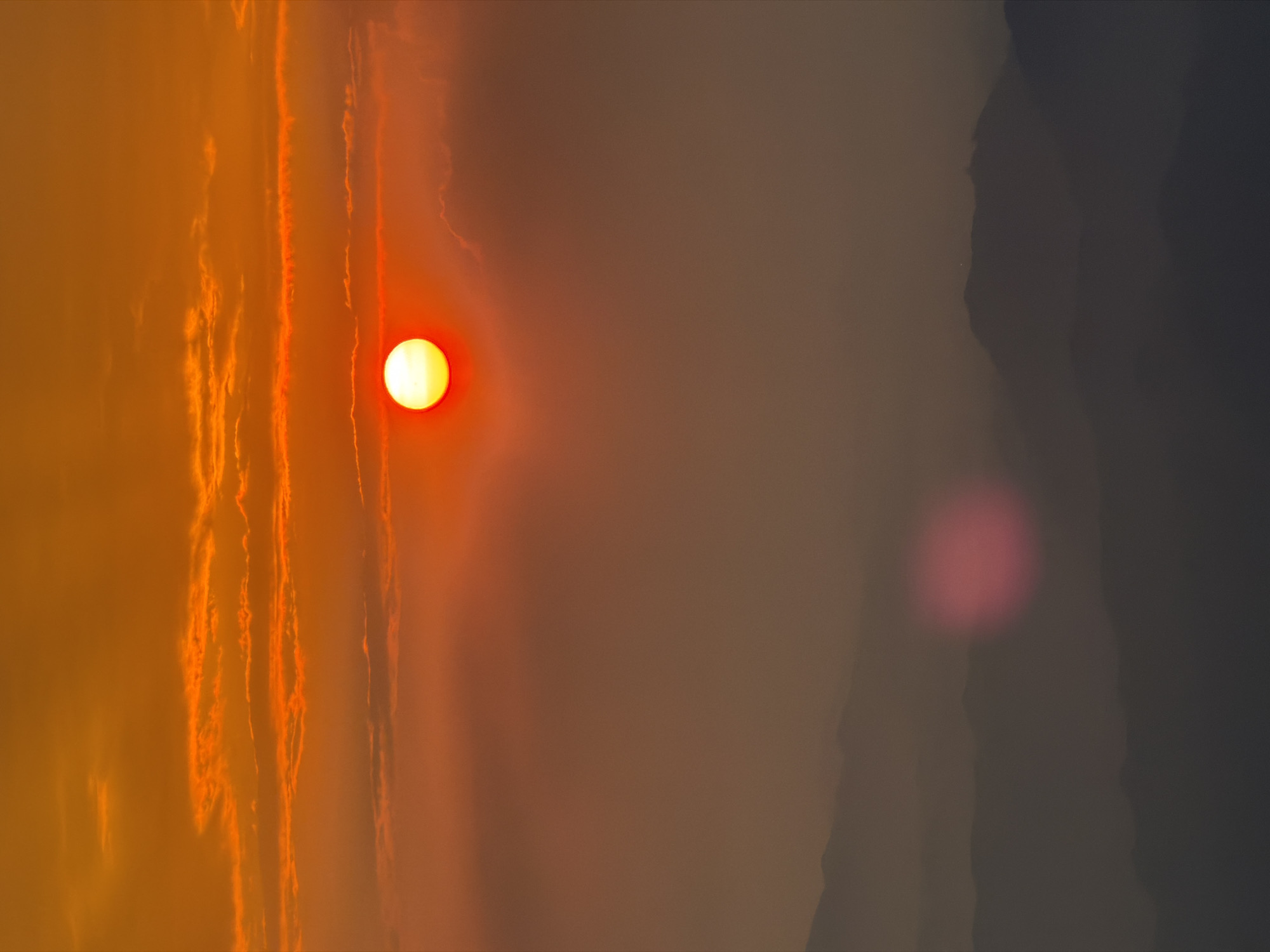 The original image presents a striking, vertically oriented view of a sunset or sunrise over a hazy landscape, characterized by warm, intense colors transitioning to cooler, darker tones. On the left side of the frame, a bright, circular yellow sun, surrounded by a red glow, is the focal point, casting its light through a thick atmosphere. Adjacent to the sun, the sky is filled with prominent vertical streaks and patches of orange and red, indicative of clouds or dense atmospheric haze illuminated by the low sun. Towards the right, the scene transitions dramatically into a darker, more muted area of deep grey and brown, which appears to be a silhouette of distant mountains or hills shrouded in heavy haze or shadow. A blurred, circular red-pink light anomaly, possibly a lens flare, is visible in the lower right portion of the image. The overall impression is a powerful, dramatic, and potentially smoky or polluted sky at dawn or dusk, possibly within the Baiyun District of China. There is no visible text in the image.