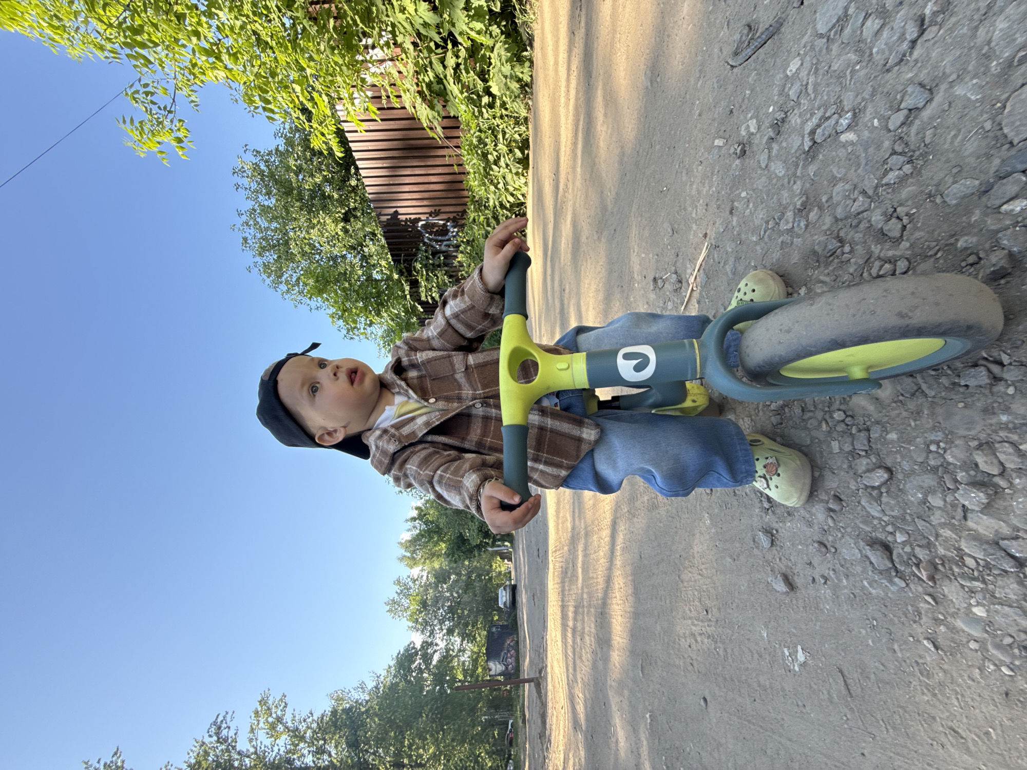 A child, estimated to be between 2 and 4 years old, is seen on a balance bike on a cracked, uneven asphalt or concrete surface interspersed with loose gravel and small stones. The child wears blue jeans, a brown plaid shirt over a yellow undershirt, a dark-colored baseball cap, and light green shoes. The bike has a lime green and gray frame. The location appears to be outdoors, likely a street or driveway with greenery visible, including trees and a fence. Clear blue sky is visible in the background. Based on the location in the city of Rusanovo, Russia, it is likely a residential area.