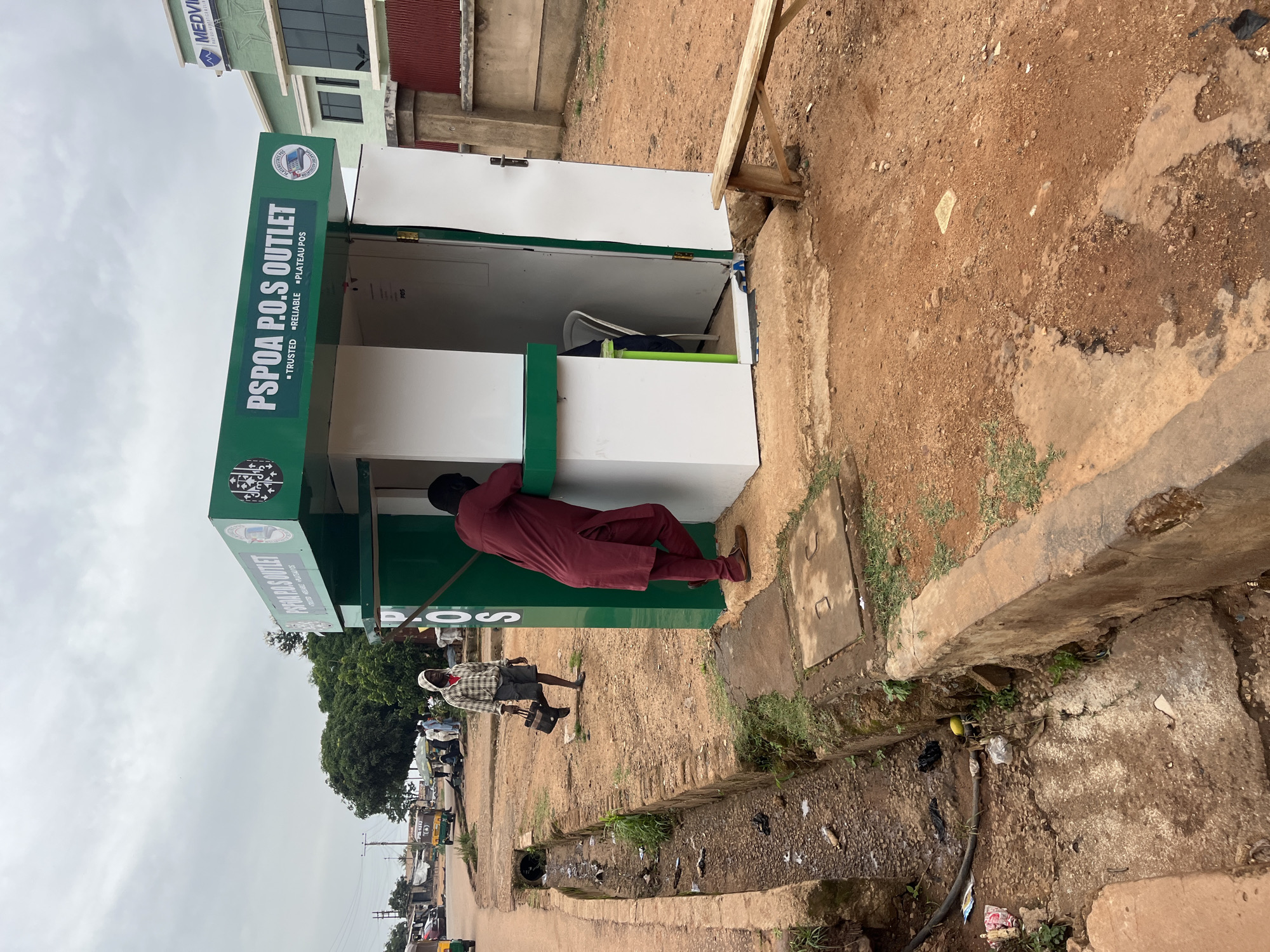 The image depicts a street scene in Bukuru, Nigeria, focusing on a small, green and white kiosk labeled "PSPOA P.O.S OUTLET". A man in a maroon tunic and pants is leaning into the kiosk, appearing to interact with someone or something inside. Another man, wearing a plaid hoodie, shorts, and a checkered shirt, walks away from the kiosk, carrying a black bag. The kiosk is situated on a dirt and concrete path, with a drainage ditch running alongside it. In the background, there are more people, some motor vehicles, and trees. A multi-story building with a "MEDVIEW" sign is visible in the distance. The sky is overcast.