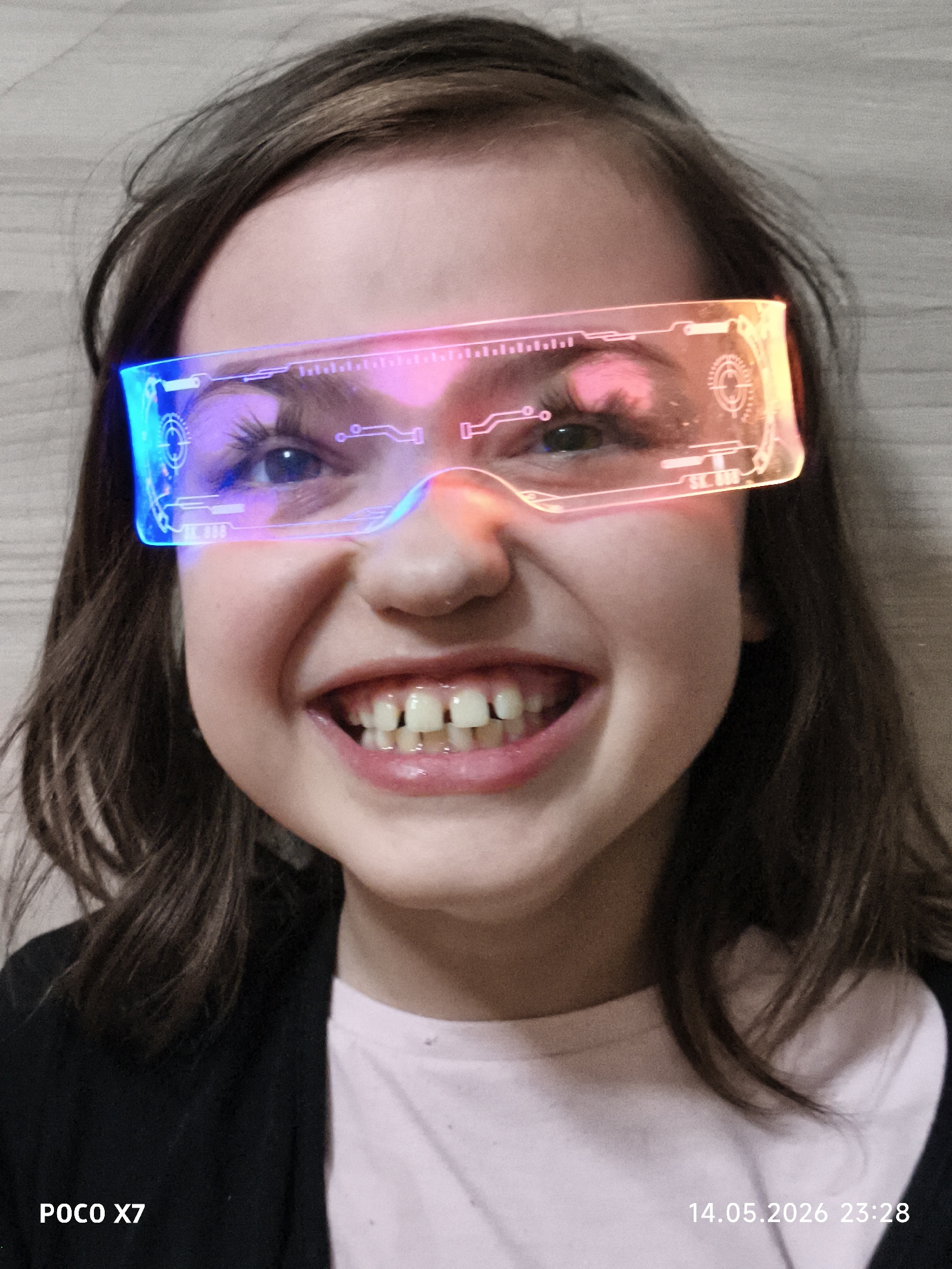 A young girl is centered in the frame, wearing futuristic-looking, frameless glasses that glow with neon colors. The glasses have a distinct blue hue on the left, transitioning through pink in the center, to a reddish-orange on the right. They feature electronic circuit patterns and numerical displays embedded within the lenses. The girl is smiling broadly, revealing her teeth. She has brown hair and is wearing a light pink t-shirt with a dark jacket over it. The background appears to be a neutral-toned wall. The text "POCO X7" is located in the bottom left corner of the image, and "14.05.2026 23:28" is in the bottom right corner. No location details are discernable.