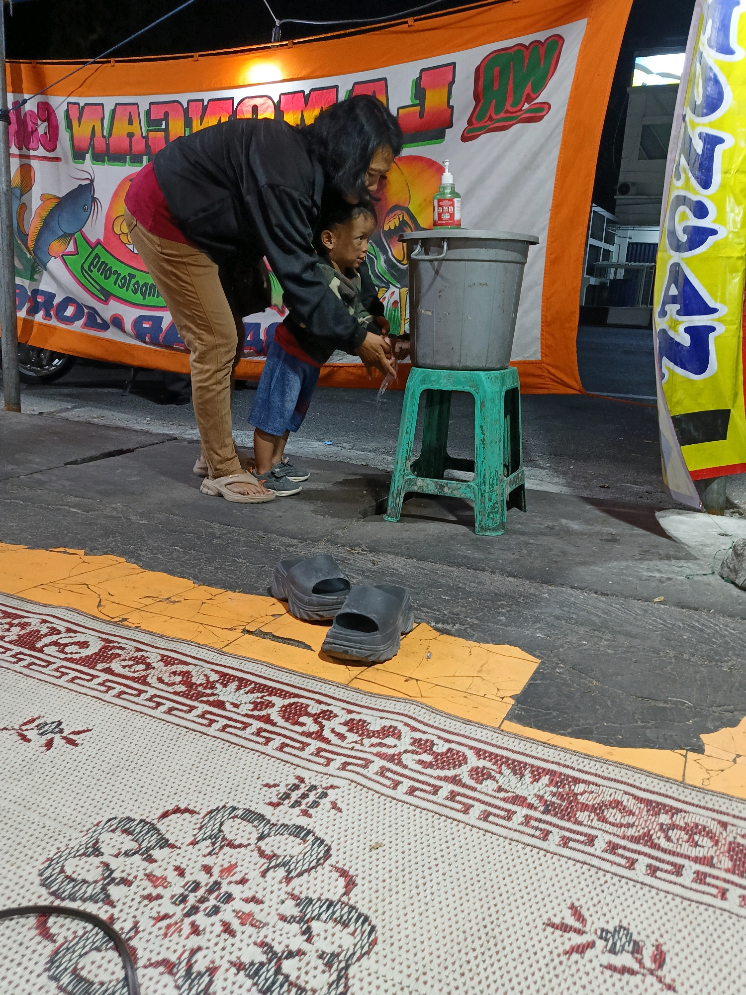 The image depicts a mother and child standing on a paved area, likely outdoors, with a street and a building visible in the background. A large banner with colorful text and graphics, possibly advertising a business, hangs behind them. In the foreground, a woven mat is spread on the ground, and a pair of flip-flops lies nearby. The mother, wearing a black jacket and tan pants, is bent over, assisting a young child in washing their hands. The child, dressed in a camouflage jacket and blue shorts, is reaching towards a grey bucket placed on a green plastic stool. A bottle of hand sanitizer is visible on top of the bucket. The scene appears to be in Kebondalem Kidul, Indonesia, as indicated by the street signage. The lighting suggests it is either dusk or nighttime, with artificial lights illuminating the scene.