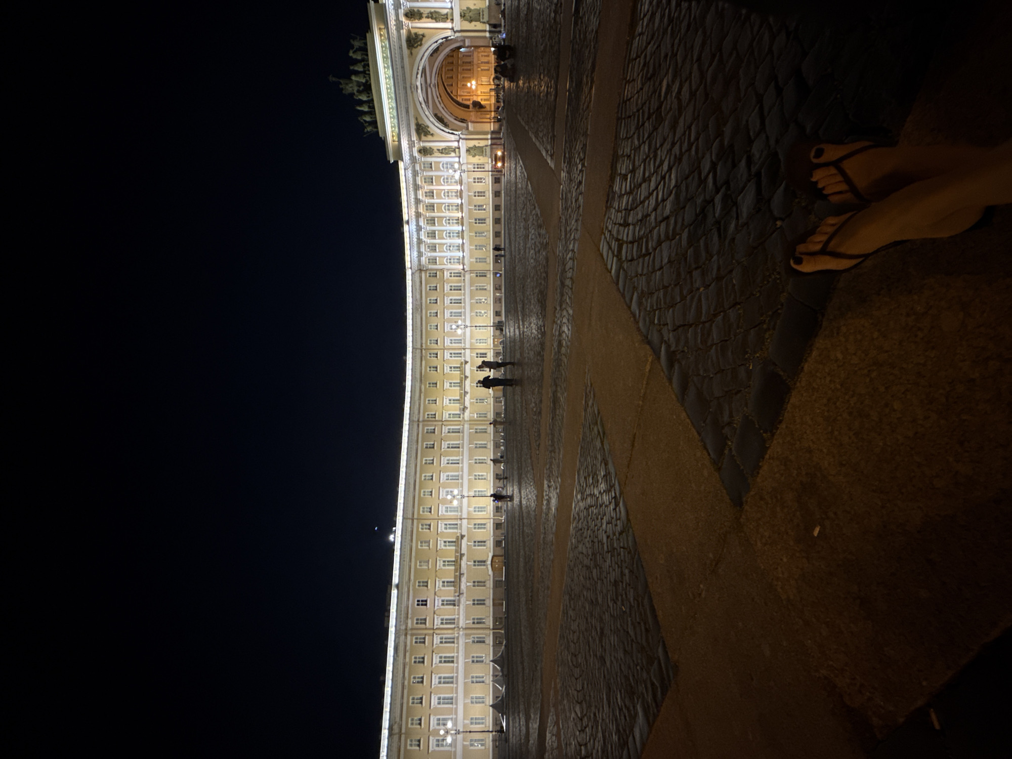 A low-angle night shot captures the grandeur of the General Staff Building and the Triumphal Arch at Palace Square in Saint Petersburg, Russia. The neoclassical building, illuminated by strings of lights along its facade and warm light spilling from interior windows, curves around the vast, cobblestone expanse of the square. The Triumphal Arch, adorned with sculptures and crowning chariots, stands prominently on the right, framing a view into the interior courtyard of the building complex, where more lights are visible.
Several individuals are scattered across the square. Two figures stand near the building on the left, and a group of people is seated or reclining on the ground beneath the arch on the right. In the foreground, the feet of the photographer are visible, clad in black flip-flops. The cobblestone paving of the square is wet, reflecting the artificial lights, suggesting recent rain or that the area has been recently washed. The night sky is dark and clear, with a faint, distant light, possibly a planet or a distant aircraft, visible near the top center of the frame. The overall atmosphere is serene and majestic, a typical scene of a historic city square at night.