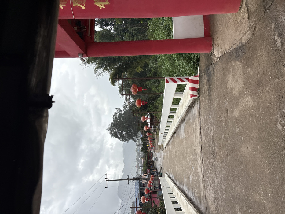 An image captures a wet, concrete road and bridge in Ban Mai Saha Sampan, Thailand, on an overcast day. The perspective is from beneath a dark, indistinct overhead structure, possibly the interior roofline of a vehicle.
In the immediate foreground, a rough concrete road surface is visible, leading to a concrete bridge. On the right, a red and white striped barrier marks the approach to the bridge, adjacent to a large, red archway or gate structure with gold-colored script on its visible column. Lush green vegetation grows densely on the right side beyond this archway.
The bridge features white concrete balustrades along both sides. The road continues slightly uphill beyond the bridge, narrowing into the distance. Numerous red, spherical Chinese lanterns hang from dark poles that line both sides of the road and bridge, extending into the background.
A white sedan is visible on the road in the mid-distance, positioned centrally. Further in the background, a cluster of residential or commercial buildings with varied roofs lines the road. Utility poles with overhead wires are present. Distant mountains, partially obscured by atmospheric haze or clouds, form the horizon under a heavily clouded sky. No individuals are visible in the scene.