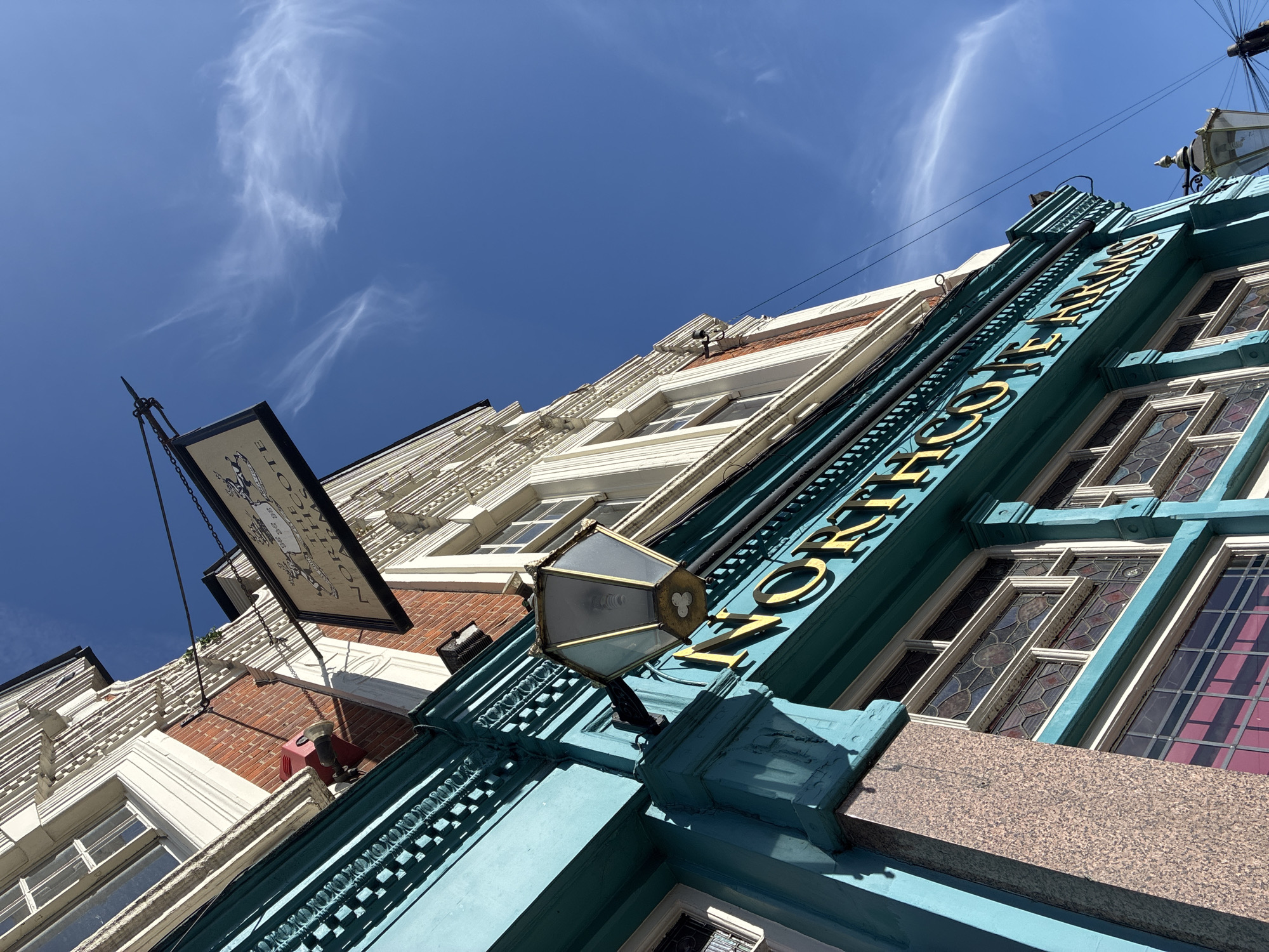The image shows a low-angle view of the architecture in London, UK. The main focus is the building of The Northcote Arms. The name "NORTHCOTE ARMS" is displayed vertically on the side of the building in gold lettering against a turquoise background. An ornate, dark-colored sign hangs on the left, attached to the building via chains. The sky above is a clear blue with wispy white clouds. Architectural details of adjacent buildings are visible.