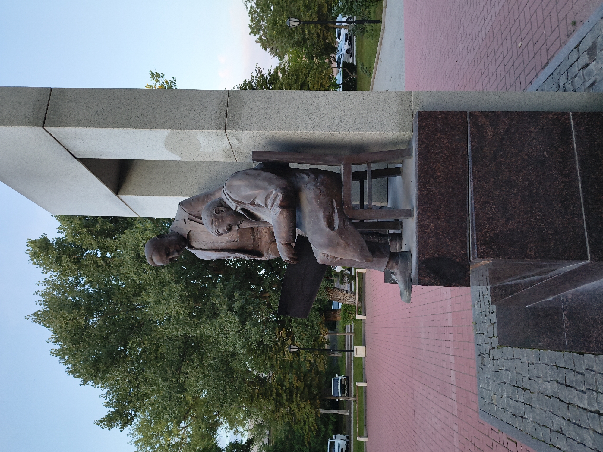 The image shows a bronze sculpture of two men, one seated and one standing, in a city park. The standing man is looking down at something the seated man is holding, possibly a book or a document. Both men are dressed in formal attire. The sculpture is located in Mikhaylovka, Russia, on a paved area with red brick pavers. Behind the sculpture, there are trees and a modern architectural structure made of concrete. In the background, cars are parked along a street, and there are benches and lampposts. The sky is clear and blue.