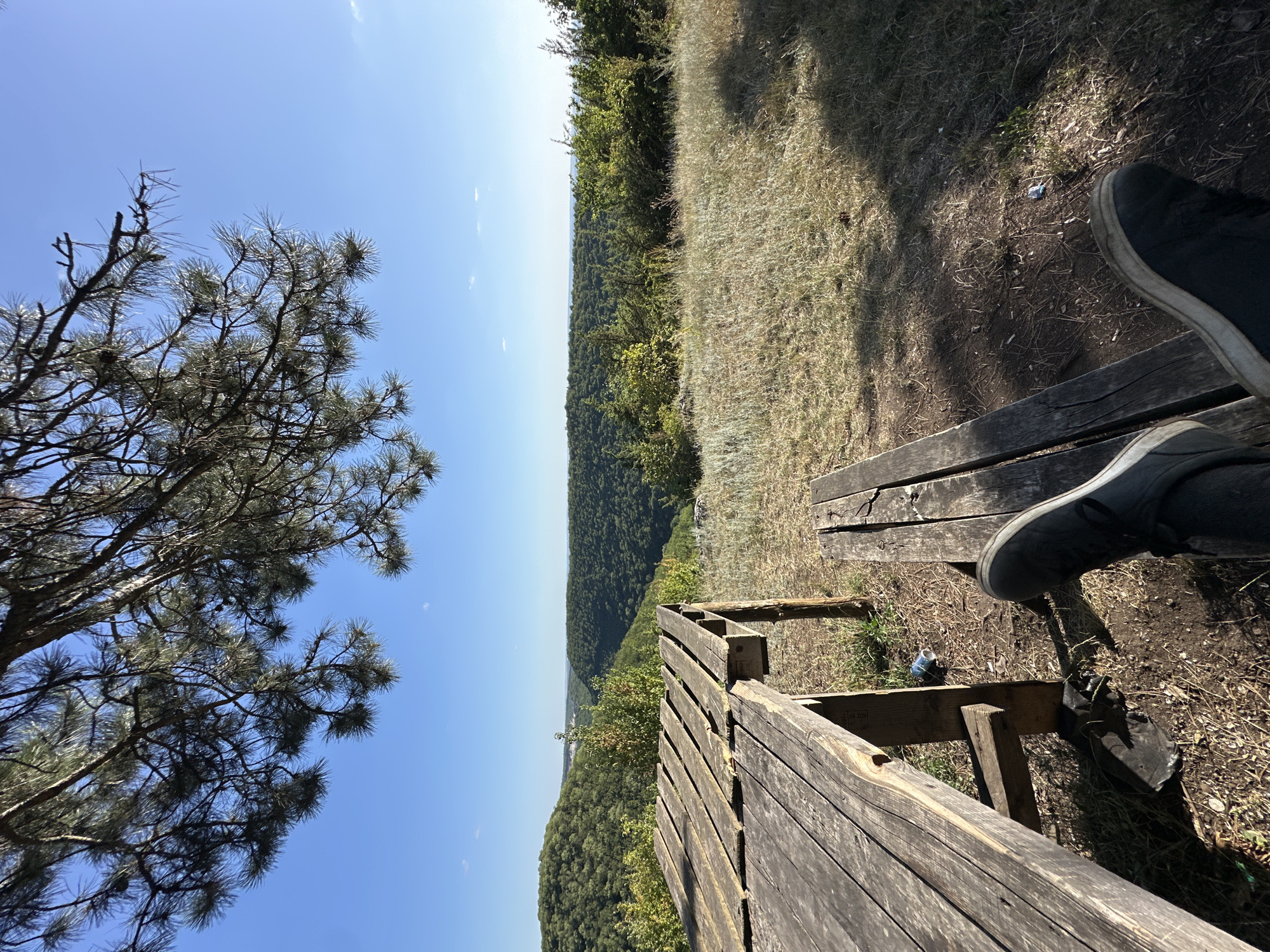 The image depicts a scenic overlook from a wooded area, likely a park or natural reserve. In the foreground, a rustic wooden picnic table and bench are positioned on a dry, grassy ground. A pair of dark sneakers is visible, suggesting someone is seated on the bench. A blue can is on the ground near the table. The background offers a panoramic view of lush green valleys and hills under a clear blue sky. The scene evokes a sense of tranquility and natural beauty. The specific location in the city of Nihyn, Ukraine, is not ascertainable from the image alone.