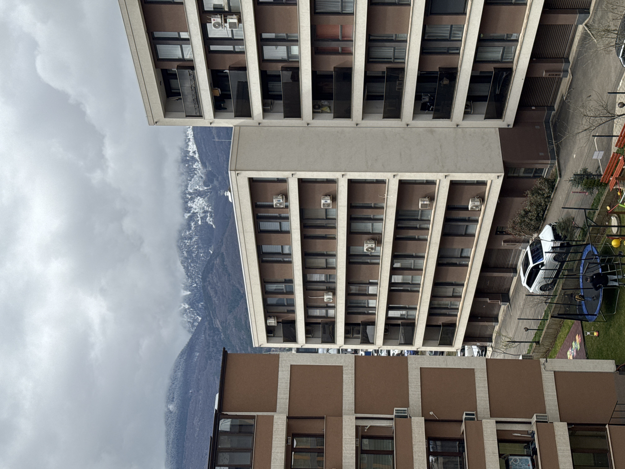 The image depicts a view of apartment buildings in what appears to be Sofia, Bulgaria. The architectural style is consistent with residential buildings constructed during the socialist era, characterized by repetitive modular design and use of concrete. Mountains are visible in the background partially obscured by clouds. In the foreground, some recreational equipment and a parked car are present. The sky is overcast.