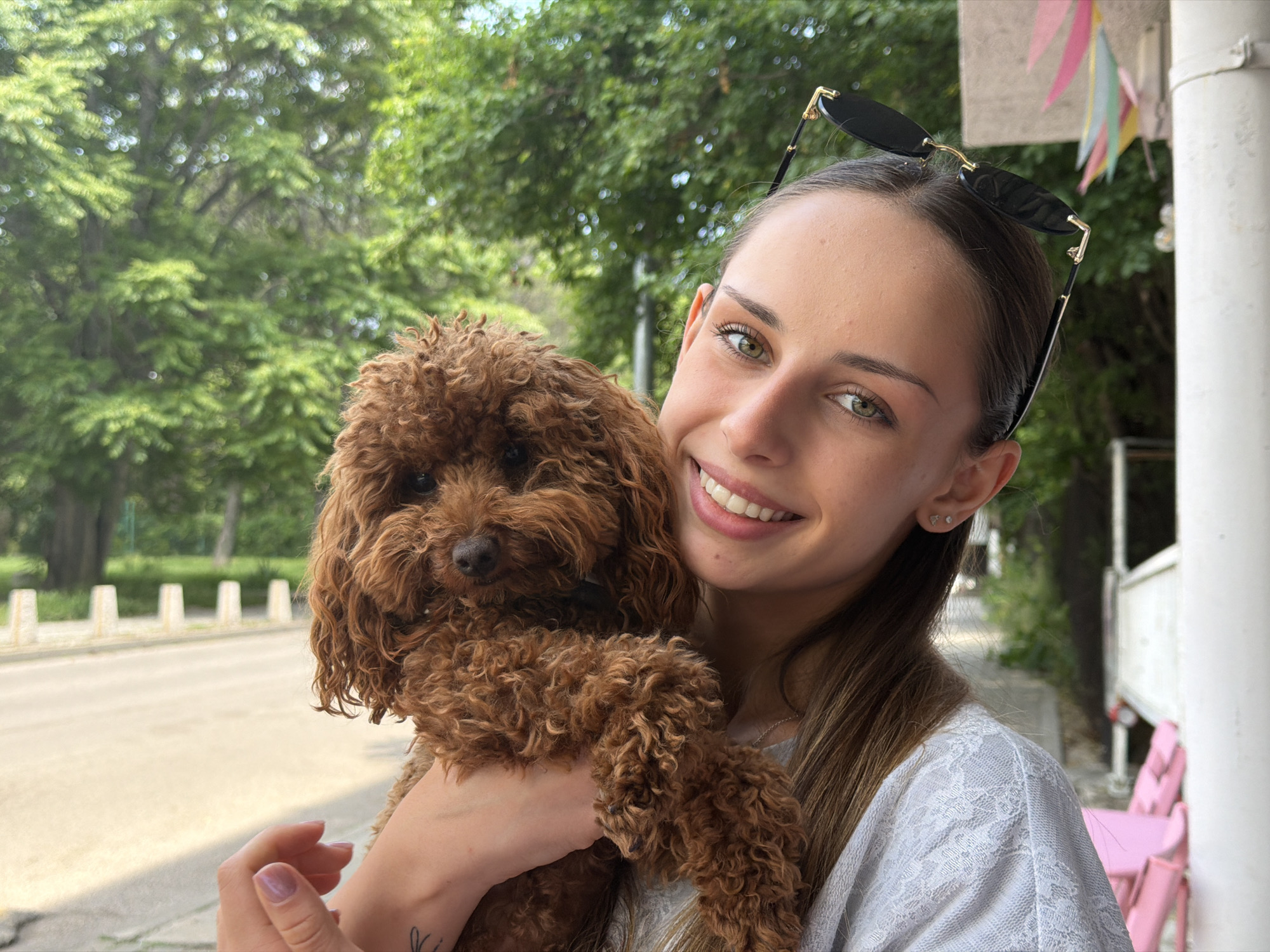 A young adult female, appearing Caucasian with medium brown hair pulled back, green eyes, and a broad smile, is depicted outdoors holding a small, reddish-brown, curly-coated dog, consistent with a miniature poodle. Black sunglasses with gold accents are perched on her head. She wears a light-colored top featuring a lace overlay pattern and has two small stud earrings in her visible ear. A faint dark tattoo is partially visible on her left forearm. The dog is positioned with its head slightly to the right, displaying dark eyes and nose. The scene is illuminated by bright daylight, creating a shallow depth of field that keeps the subjects in sharp focus. The background consists of out-of-focus lush green foliage from trees, a paved area with white bollards, and the right side of a white building featuring a prominent white column. Adjacent to the building, pink outdoor chairs and a table are visible, with colorful triangular bunting strung above, indicating an outdoor cafe or leisure area. The specific address in Sofia, Bulgaria, is not identifiable from the visible cues, which depict a generic urban park and outdoor cafe setting.