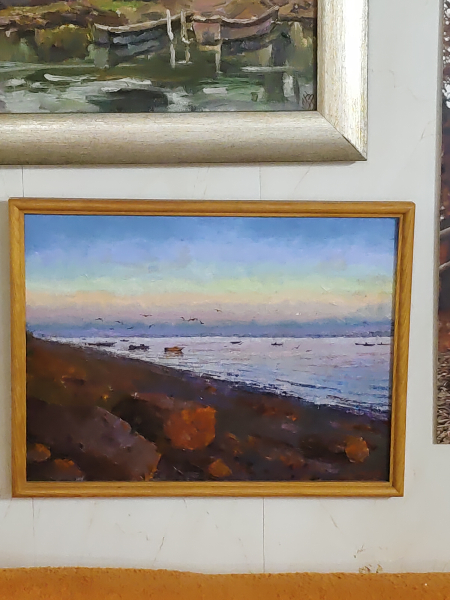 A framed oil painting depicts a serene seascape at sunset. The foreground is a dark, rocky shore, with patches of vibrant orange. The sea stretches out to the horizon, reflecting the soft hues of the sky, transitioning from pale blue at the top to soft pinks and yellows near the horizon. Several small boats are scattered on the water, and a flock of birds flies in the distance. The painting is displayed on a white wall above an orange upholstered object. The location is Olginskaya, Russia.