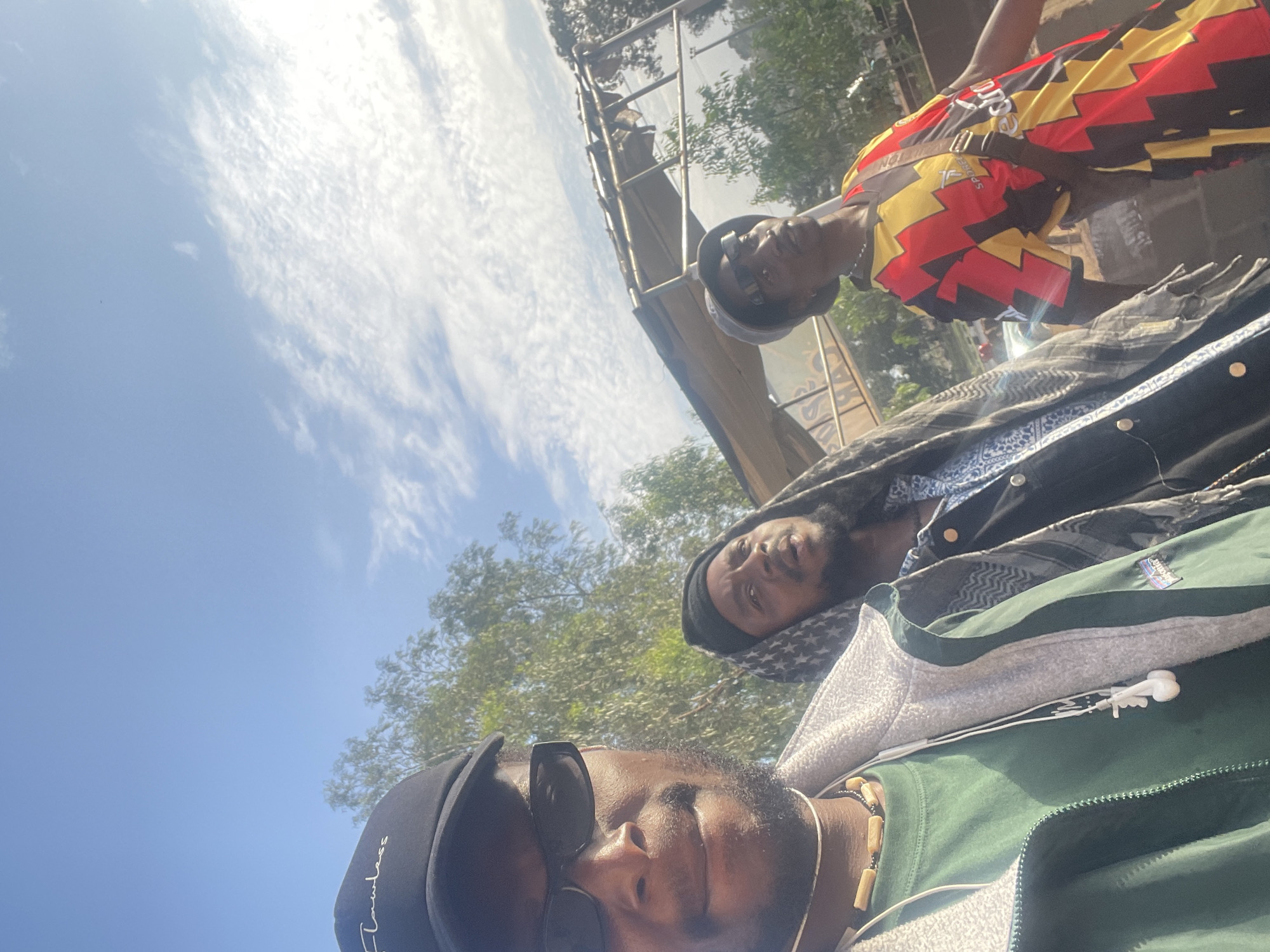 Three men are visible in the image, oriented at an angle towards the sky. One man in the foreground wears sunglasses and a black baseball cap. A second man is positioned above the first, wearing a black headdress with white star and moon patterns. A third man stands above the second, clad in a brightly colored shirt featuring geometric patterns. The setting appears to be outdoors, with trees and the sky visible in the background. An indistinct metal structure is partially visible above the third man. The location is Bar Jirgi Summa, Nigeria, however the specifics of the environment or the activities of the people cannot be determined from the image.