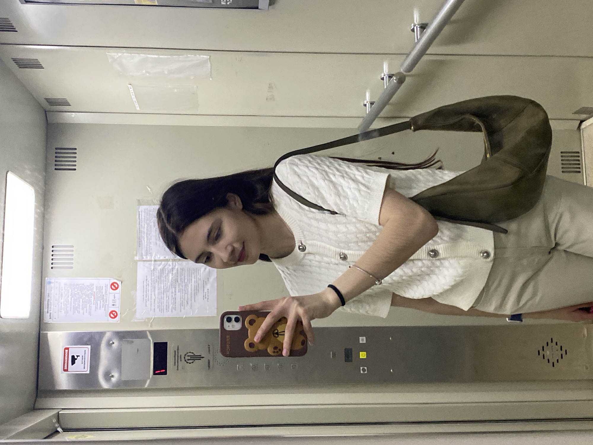 A young woman is taking a selfie in an elevator in Votkinsk, Russia. She is holding her phone, which has a brown case with a teddy bear design, up to the mirror to capture the image. The woman is wearing a white cardigan with decorative buttons, light-colored pants, and has a dark-colored shoulder bag. The elevator has a metal control panel with buttons and a digital display indicating the current floor, which is labeled as "1". Various safety and information signs are posted on the elevator walls. A handrail is visible in the background. The interior of the elevator is utilitarian, with plain walls and minimal decoration.