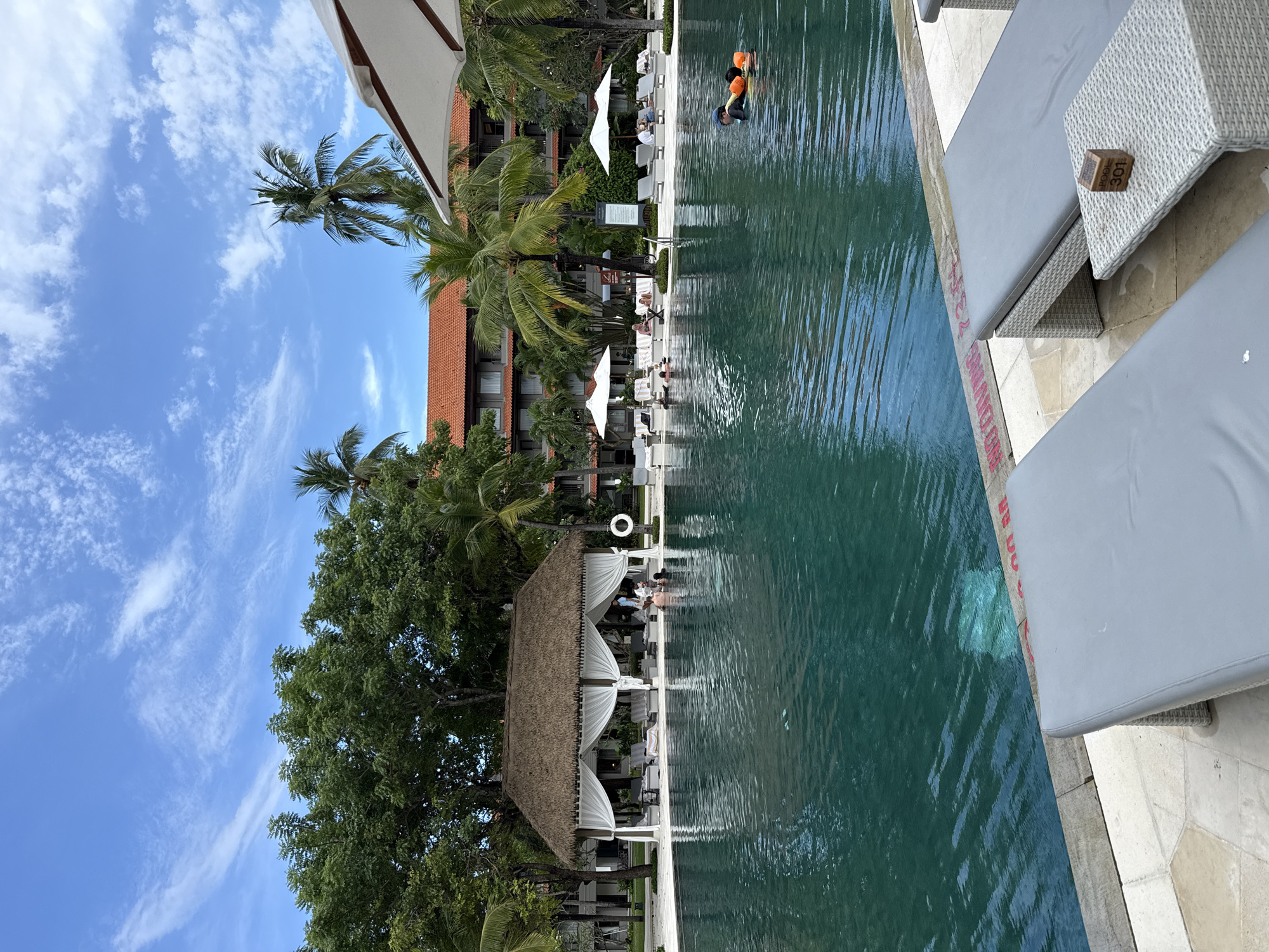 The image depicts a tranquil resort scene in Nusa Dua, Indonesia, showcasing a large swimming pool adjacent to a series of lounge chairs and poolside cabanas. The pool water reflects the bright sky and nearby buildings, while two individuals, wearing snorkeling gear, are visible in the water.
The scene is characterized by palm trees, a building with a red tiled roof, and a blue sky with wispy clouds. In the foreground, there are white lounge chairs with gray cushions. Small wooden signs are on the tables next to the lounge chairs. The poolside cabanas have thatched roofs and white curtains. The overall environment suggests a luxurious and relaxing vacation setting.