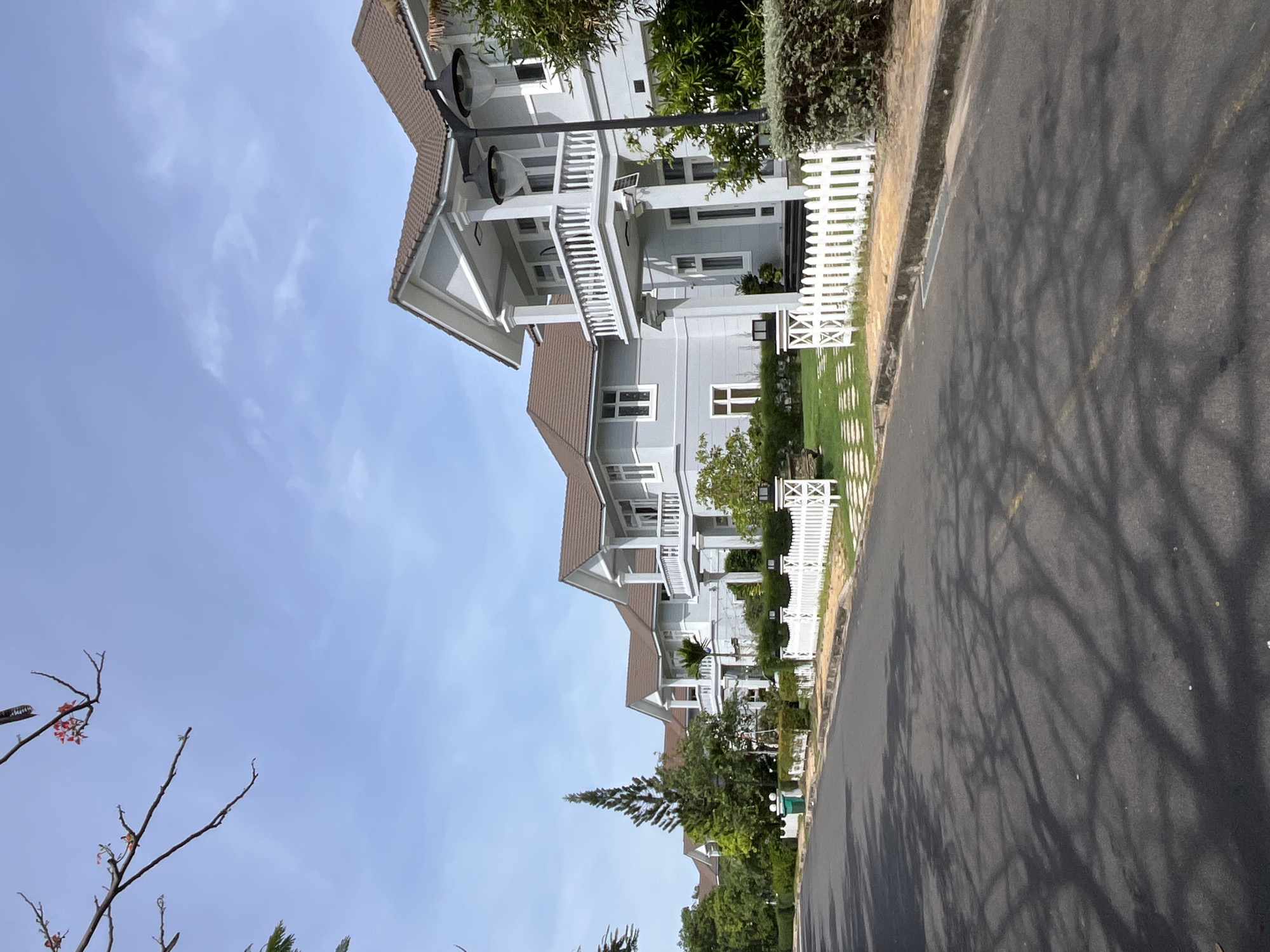 The image shows a residential street lined with modern houses in Phu Thuy Ward, Vietnam. The houses are two stories tall, with light blue siding, brown tiled roofs, and white balconies supported by columns. A white picket fence runs along the front of the properties, separating them from the street. Lush green trees and well-maintained lawns surround the houses. The road is paved and appears to be in good condition, with yellow lane markings visible. Sunlight casts the shadows of trees onto the asphalt. The sky is a clear blue with a few scattered clouds. There are no people visible in the image. The scene depicts a peaceful and orderly suburban environment.