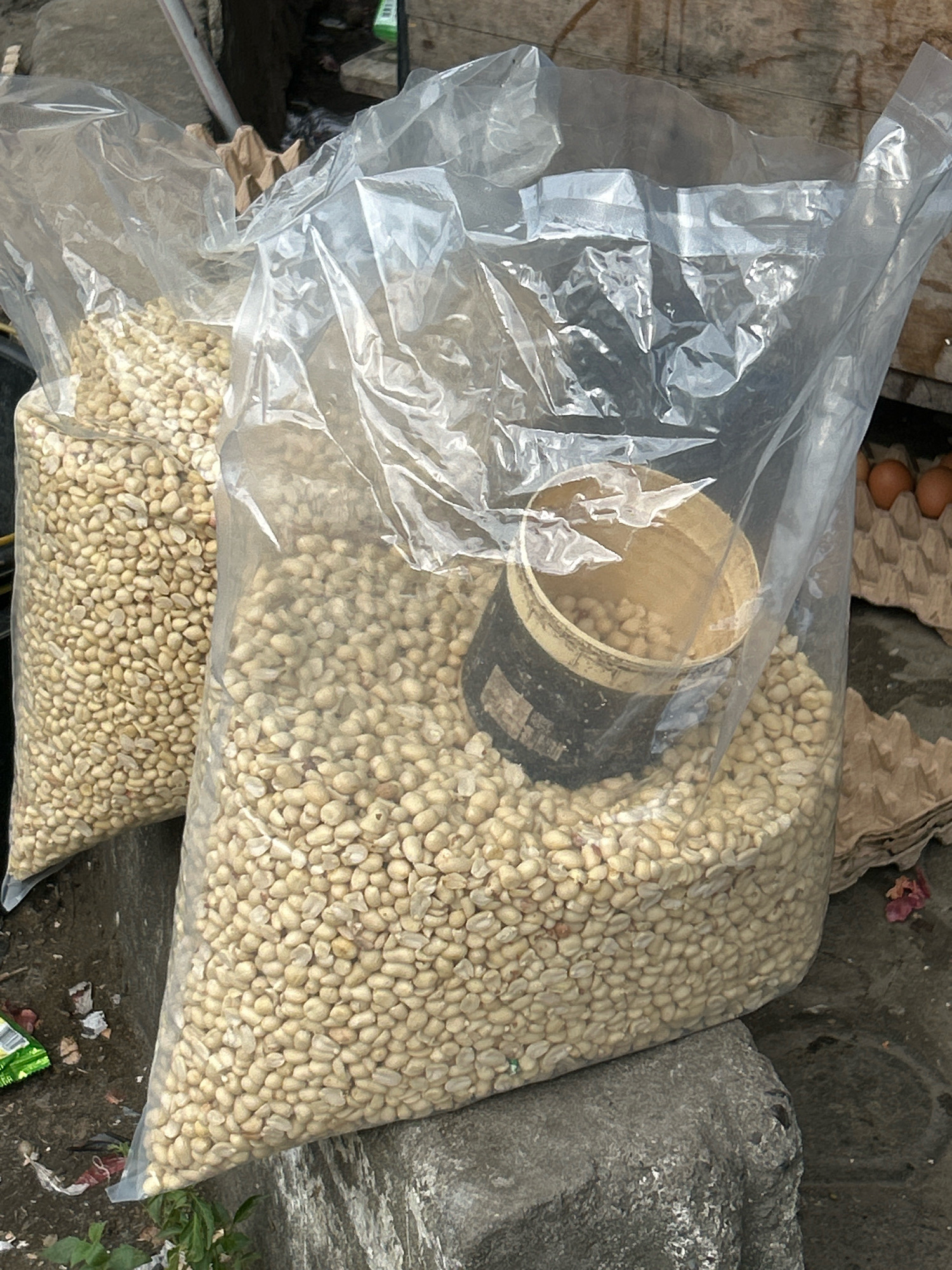 Two large, clear plastic bags filled with small, off-white beans are the primary subjects in this image. The bags are resting on a rough, grey concrete surface. The bag on the right is larger and more in focus, showing a brown, cylindrical container partially submerged in the beans. This container has a black band around its middle with some white lettering. To the right of the larger bag, a cardboard egg carton holds several brown eggs. The background is blurred, but suggests a rustic outdoor setting with wooden surfaces and some debris on the ground, including crumpled brown paper and a small green packet. A hint of green foliage is visible in the lower left corner. The scene appears to be a market stall, potentially in Banda Aceh, Indonesia, given the context provided.