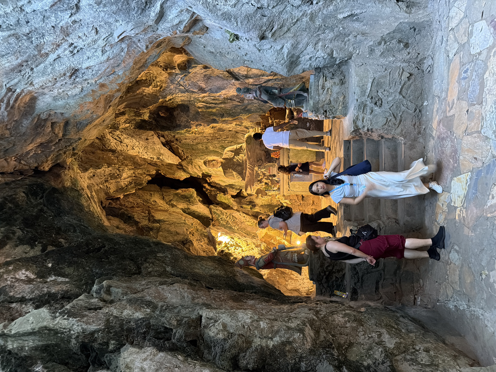 The image depicts the interior of a cave at the Marble Mountains in Đà Nẵng, Vietnam. In the background, a large, carved Buddha statue is illuminated by warm lighting, seated within the cave's natural rock formations. To the left of the Buddha, a statue of a warrior stands guard. Several tourists are visible throughout the cave. In the foreground, a woman in a light-colored dress walks down a stone staircase, carrying a dark blue bag over her shoulder. To her left, another woman with dark hair and red shorts is looking away from the camera. Further back, a man in a white shirt and beige pants stands near the warrior statue, and a woman in a gray shirt with a backpack is walking away. A small child in a dark dress is also present. The cave walls are textured and rough, with natural rock formations and some foliage. The floor is paved with irregular stones. The lighting creates a dramatic contrast between the brightly lit Buddha and statues and the darker, shadowed areas of the cave.