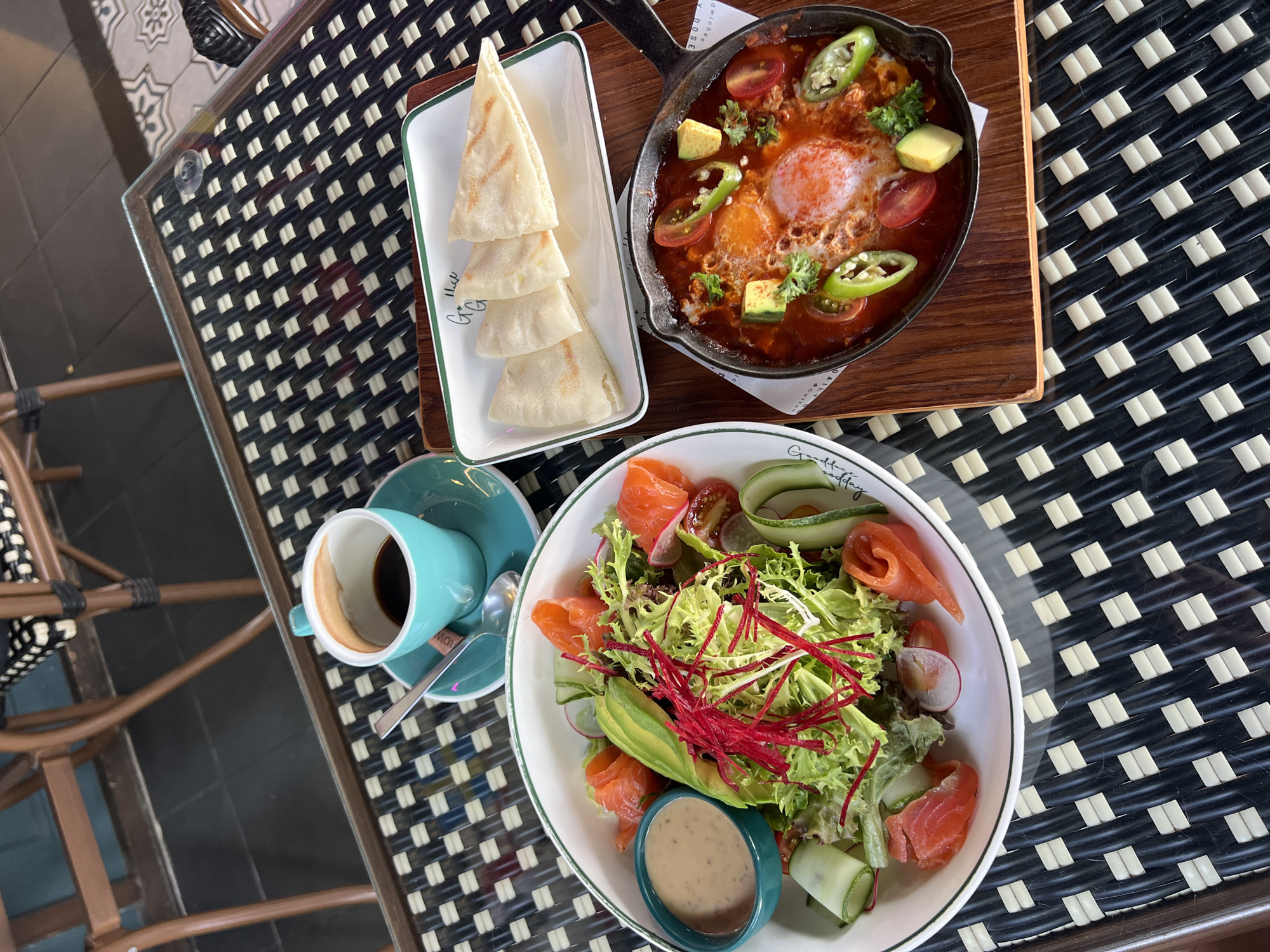 The image shows a meal laid out on a black and white woven table in what appears to be an outdoor seating area at a restaurant in Choeng Thale, Thailand. On the left is a white bowl filled with a salmon salad, including mixed greens, avocado slices, cucumber, cherry tomatoes, and shredded red vegetables. A small teal bowl of salad dressing sits to the left of the salad. In the center of the table is a teal cup filled with dark coffee, resting on a matching saucer, with a spoon. To the right of the coffee is a rectangular white plate holding four triangular pieces of flatbread. Below the flatbread is a black cast-iron skillet containing shakshuka, a dish with eggs poached in a tomato and pepper sauce, garnished with chopped herbs, avocado, and sliced jalapeños. The background shows parts of outdoor cafe chairs with wooden frames and woven seats. The table itself has a glass top with a slightly reflective surface.