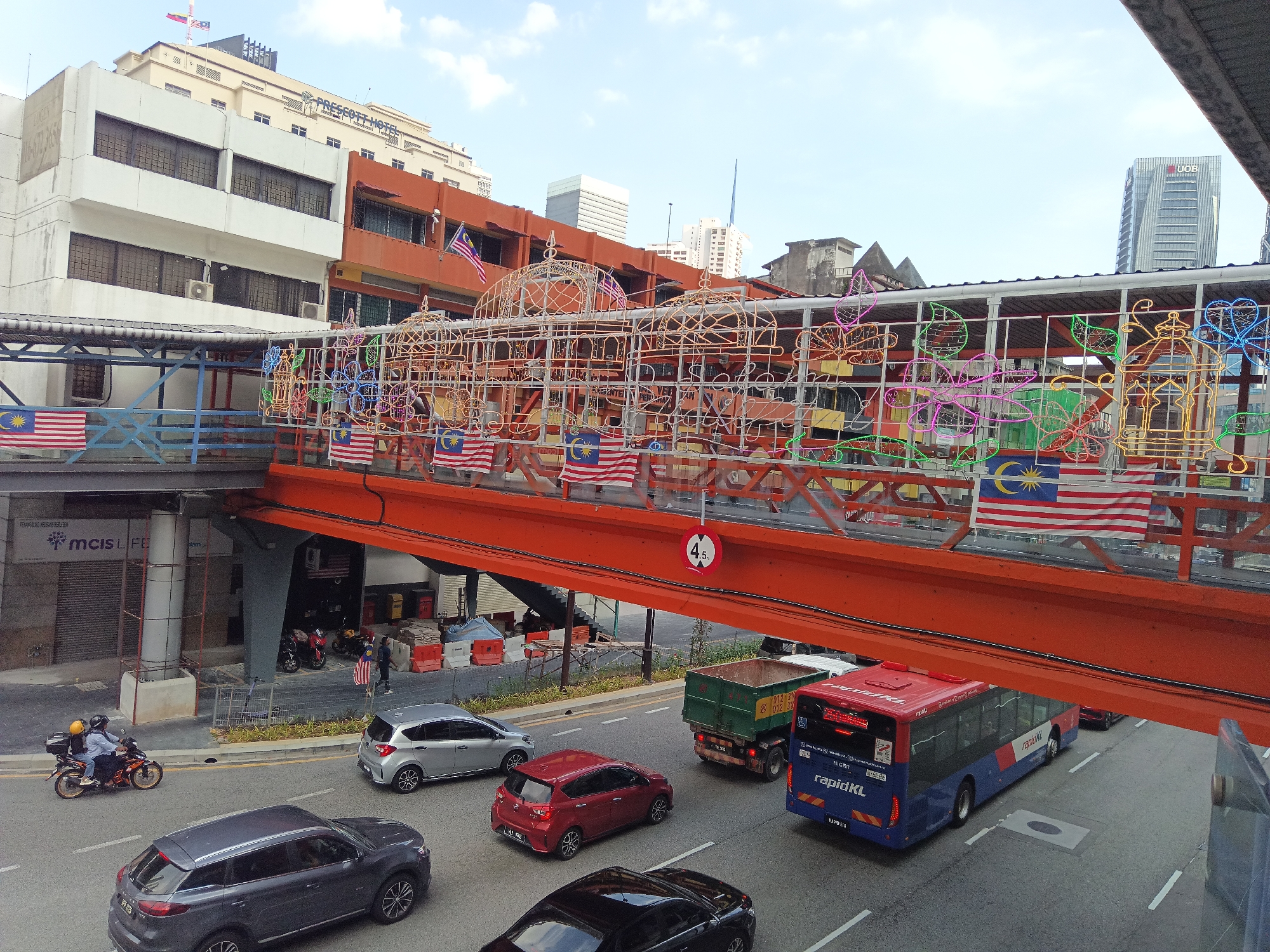 The image depicts an elevated orange pedestrian bridge adorned with decorative lights and Malaysian flags, spanning over a street in Kuala Lumpur, Malaysia. The bridge, likely a part of a commercial area or transportation hub, overlooks a busy roadway with several vehicles, including cars, a delivery truck, and a blue and red RapidKL bus. The surrounding buildings suggest an urban setting with commercial establishments and varying architectural styles. The pedestrian bridge appears to be decorated for a festive occasion, with colorful lights forming intricate patterns and motifs. The overall scene captures a dynamic urban environment with a blend of modern infrastructure and local cultural elements.