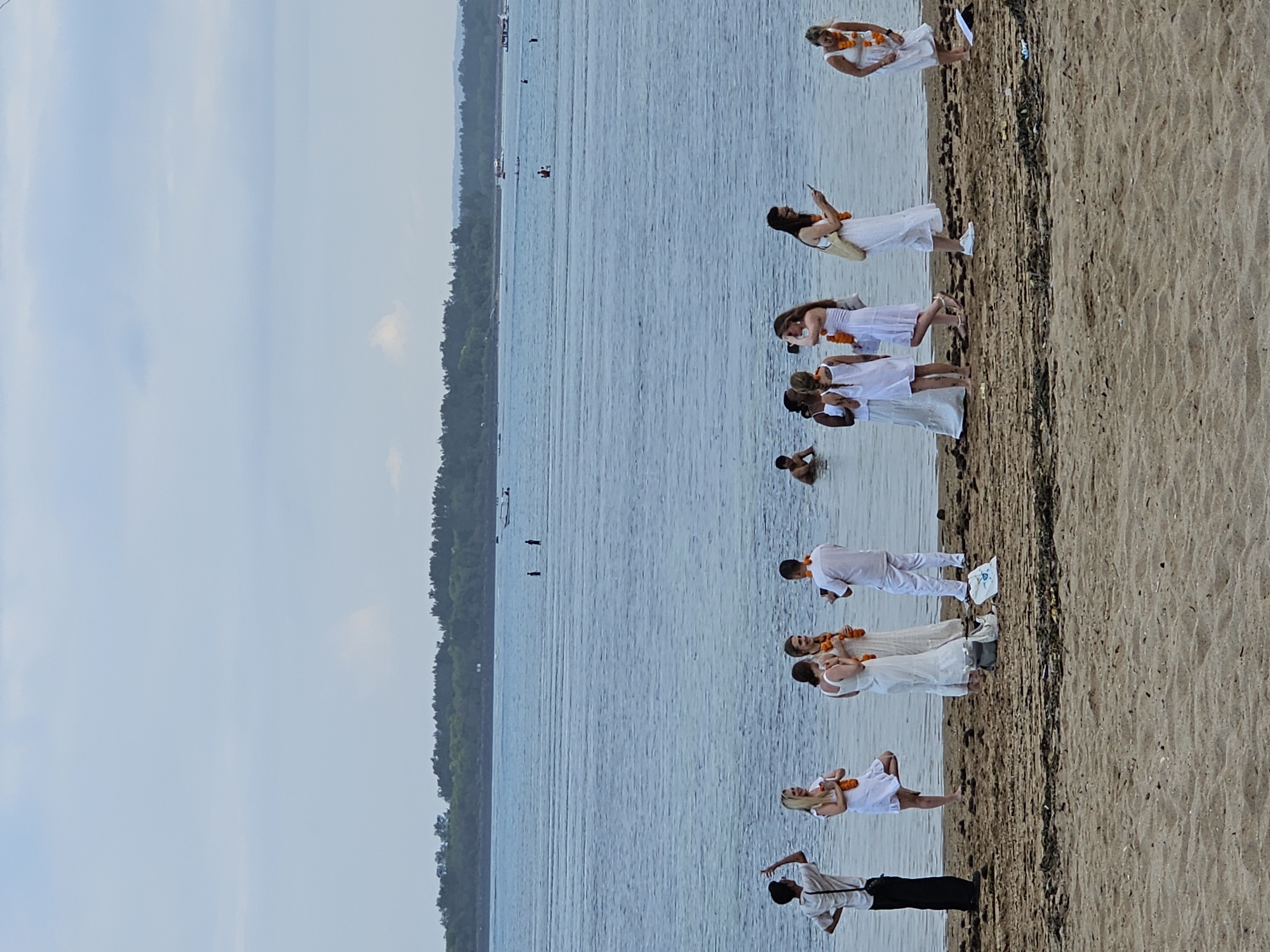 The image shows a group of people on a sandy beach next to a body of water. The sky is overcast, suggesting it might be early morning or late afternoon. The people are mostly dressed in white, with some wearing orange floral garlands around their necks, hinting at a celebratory or spiritual gathering.
Several individuals are visible on the beach, some standing and others in a kneeling or seated position. One person in the foreground, wearing black pants and a light-colored shirt, appears to be pointing. Further up the beach, a line of people in white, some with floral garlands, are arranged in a staggered formation. One woman is taking a photo with her phone. In the water, a few individuals are visible, with one person appearing to be swimming or wading.
The background shows a tree-lined shore and the calm water stretches out towards the horizon. The overall atmosphere seems peaceful and communal, possibly a yoga session, meditation retreat, or a cultural event. The location is identified as Denpasar, Indonesia, which aligns with the tropical environment suggested by the vegetation and beach setting. There is no visible text in the image.