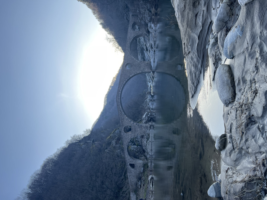 This image presents a wide-angle landscape photograph of a historic multi-arch stone bridge spanning a river within a mountainous valley. The location is near Diadovtsi, Bulgaria.
The central subject is a light grey stone arch bridge, featuring a prominent large central arch and at least three smaller arches to its left and right, all reflected clearly in the calm, dark-toned river water below. The bridge appears constructed from rough-hewn stone blocks.
In the foreground, the riverbank consists of smooth, light-colored rocks and sand, with several rounded individual stones present on the bank and partially submerged in the clear, shallow water. Sparse, dry vegetation is visible among the rocks.
Steep, tree-covered mountains flank the river valley, with the trees appearing bare or sparsely leaved, indicative of a winter or early spring season. The mountain on the left is heavily shadowed. A bright, low sun is positioned centrally in the background, directly behind the bridge, causing a significant glare against a clear, light blue sky. No individuals, vehicles, or active subjects are present in the scene.