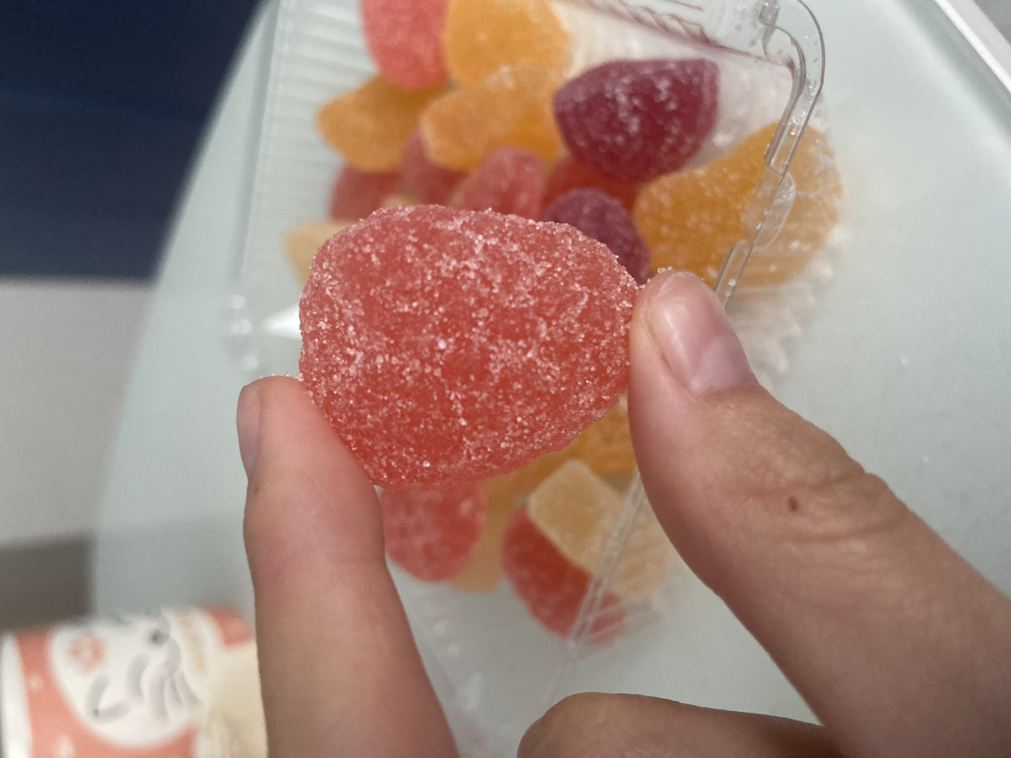 A close-up shot shows a hand holding a red, heart-shaped gummy candy coated in sugar. In the background, a clear plastic container holds a variety of colorful gummy candies in shades of red, orange, and purple. A blurry pink and white mug with a cat design is visible to the left. The scene appears to be indoors, possibly on a table, with a light blue surface in the foreground and a darker blue surface in the background. There is no information available that would allow for the determination of the location in the city of Balashikha, Russia.