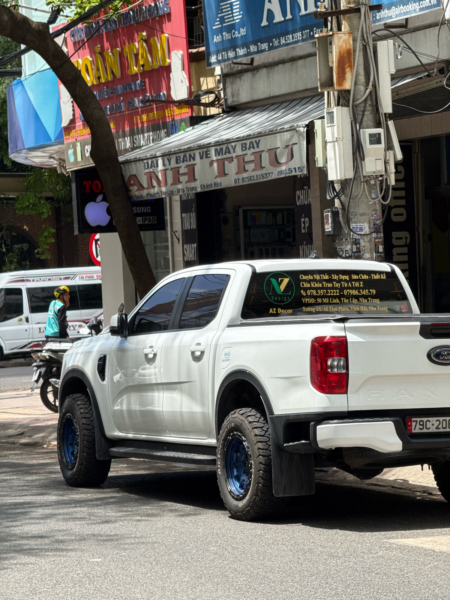 A white Ford Ranger pickup truck is parked on a street in Nha Trang, Vietnam. The truck has blue rims on its wheels and a license plate that reads "79C-208". There are several stickers on the rear window of the truck, one of which displays the text "AZ Decor". A white van is visible in the background, along with a person wearing a yellow helmet and a teal vest. The street is lined with shops and buildings, some of which have signs written in Vietnamese. Overhead, there are power lines and a tree. The scene appears to be a typical street view in a Vietnamese city.