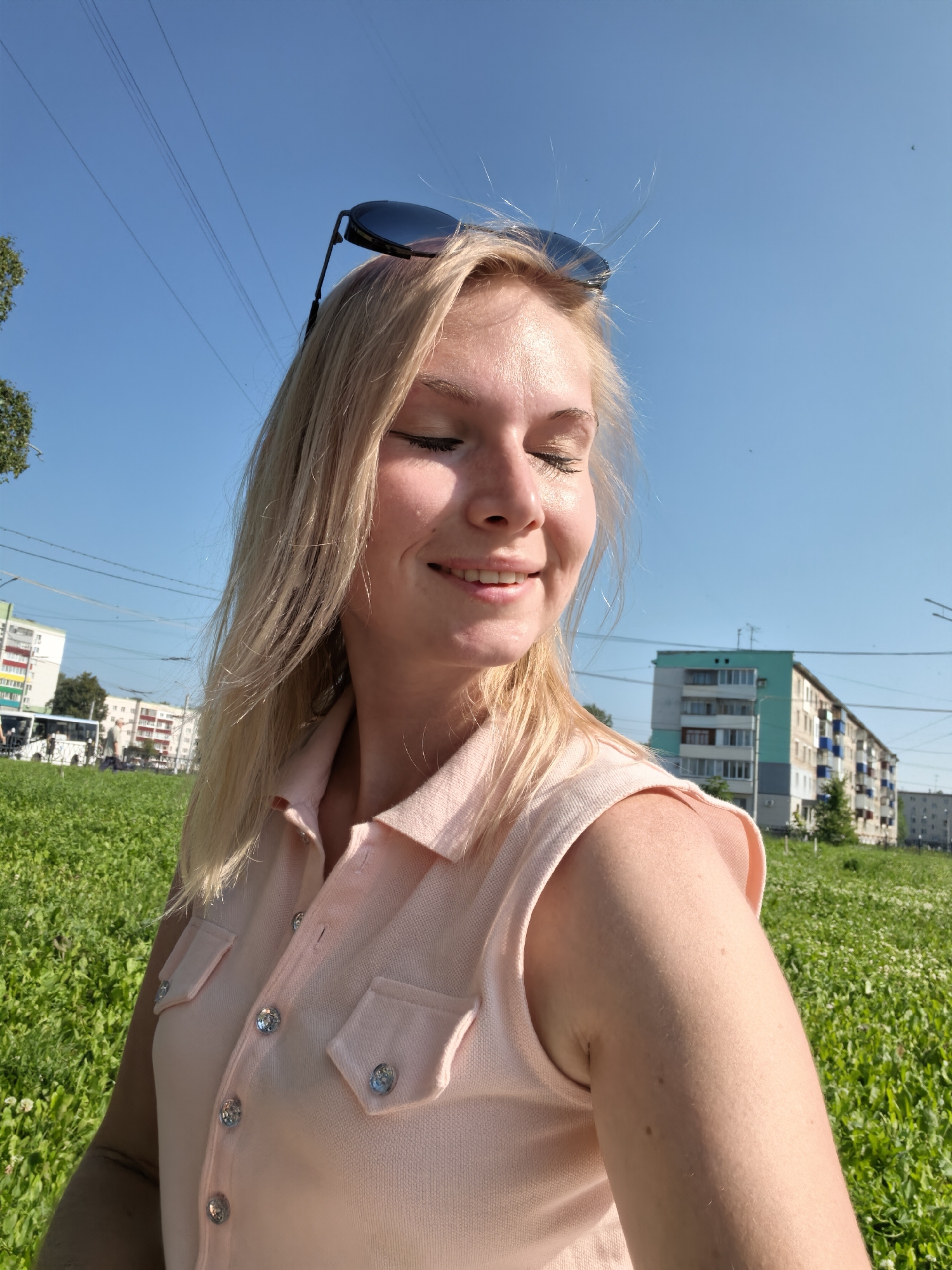 In Sterlitamak, Russia, a young Caucasian woman with blonde hair and closed eyes is captured in a close-up, low-angle selfie. She is wearing a light pink collared shirt with rhinestone buttons and two chest pockets. Sunglasses are perched on top of her head. The background features a clear blue sky with visible power lines, a patch of green grass with small white flowers, and several apartment buildings. A bus and other pedestrians are faintly visible in the distance, suggesting an urban park or street environment. The lighting indicates it is daytime, likely sunny.