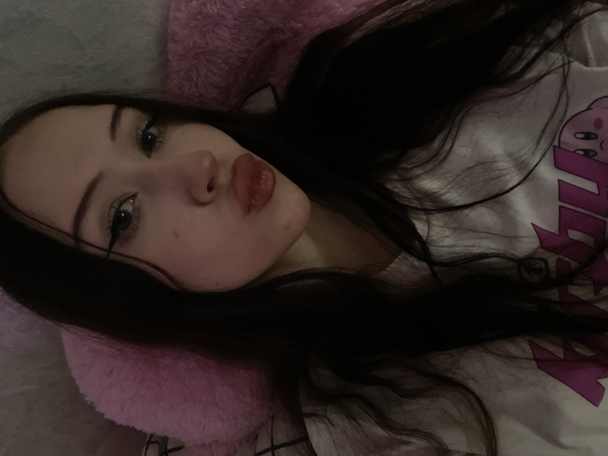 The image is a close-up selfie of a young woman with long, dark hair. She is wearing a white t-shirt with a pink graphic, possibly a character from a video game. Her lips are pursed, and she is wearing eyeliner and mascara. She is lying down, as indicated by the fluffy pink material behind her, which appears to be a pillow or a blanket. The lighting is dim, creating a soft glow on her face. The background is a blurred interior, with hints of a wall and possibly a bed. The image is taken indoors. There is no information about the location in Votkinsk, Russia.