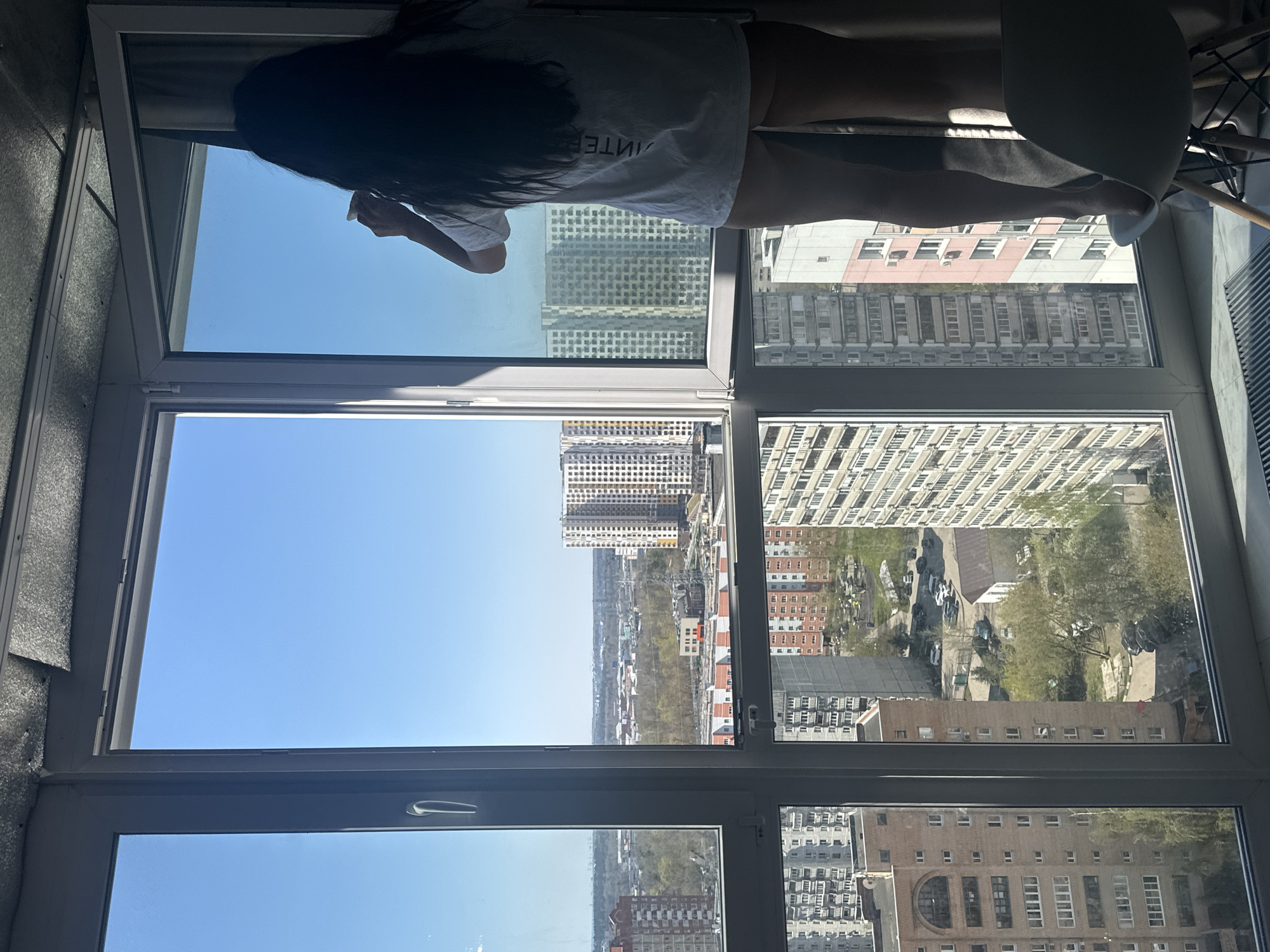 A medium, angled shot captures a person in a white t-shirt, with long dark hair, looking out of a window in a high-rise building in Отрадное, Russia. The view outside the window encompasses numerous tall buildings, a cityscape, a bright blue sky, and urban greenery. A chair is visible in the upper right corner of the frame. The window is multi-paned with a white frame. The light conditions suggest a sunny day.