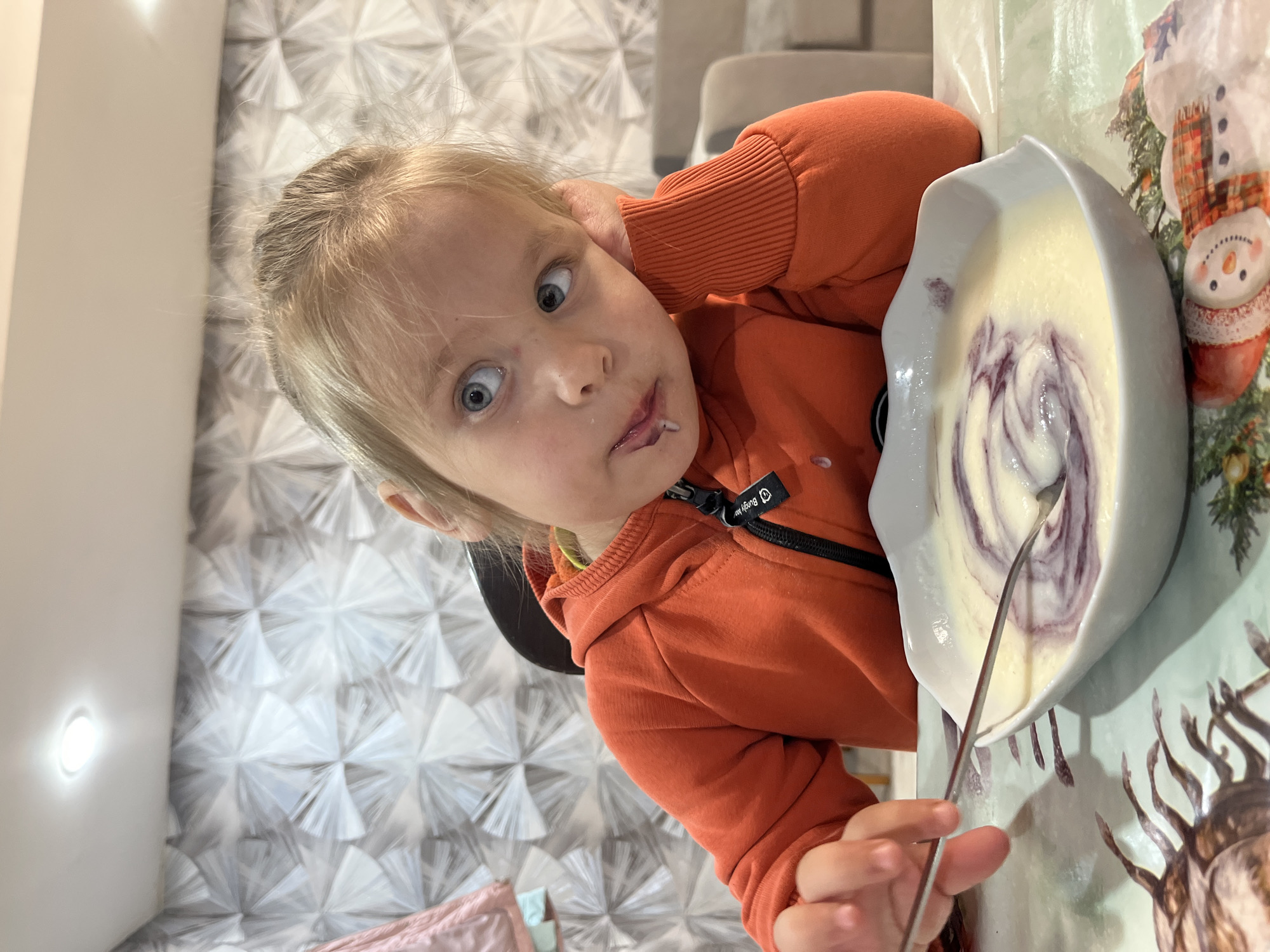 A young child with fair hair is seated at a table, angled toward the viewer, holding a spoon in one hand and a bowl containing a light-colored food with purple swirls in the other. The child's expression is wide-eyed and playful. The child wears an orange zip-up hoodie. The background consists of a wall with a patterned wallpaper and a surface that appears to be covered in a tablecloth or wrapping paper featuring various designs, including a snowman and botanical elements. The location is Chaikovsky, Russia.