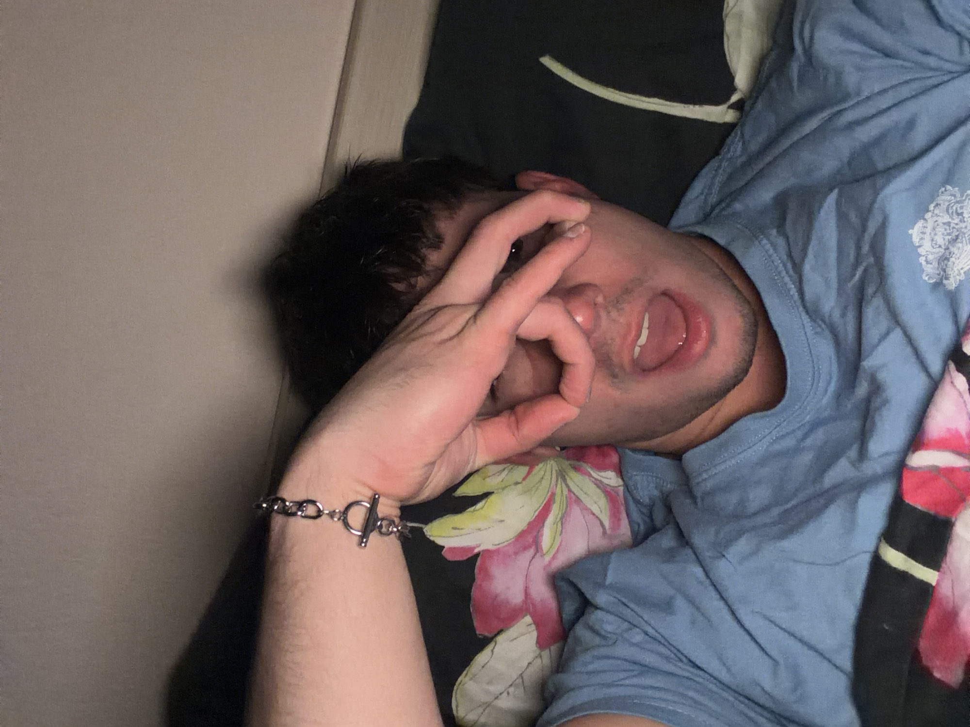 A man is lying down on a bed. He has dark hair and is wearing a light blue t-shirt. His left arm is raised, with his hand forming a circle over one of his eyes. A silver bracelet is visible on his left wrist. The bed has a dark colored sheet or blanket with a floral design. The location is not identifiable from the image.