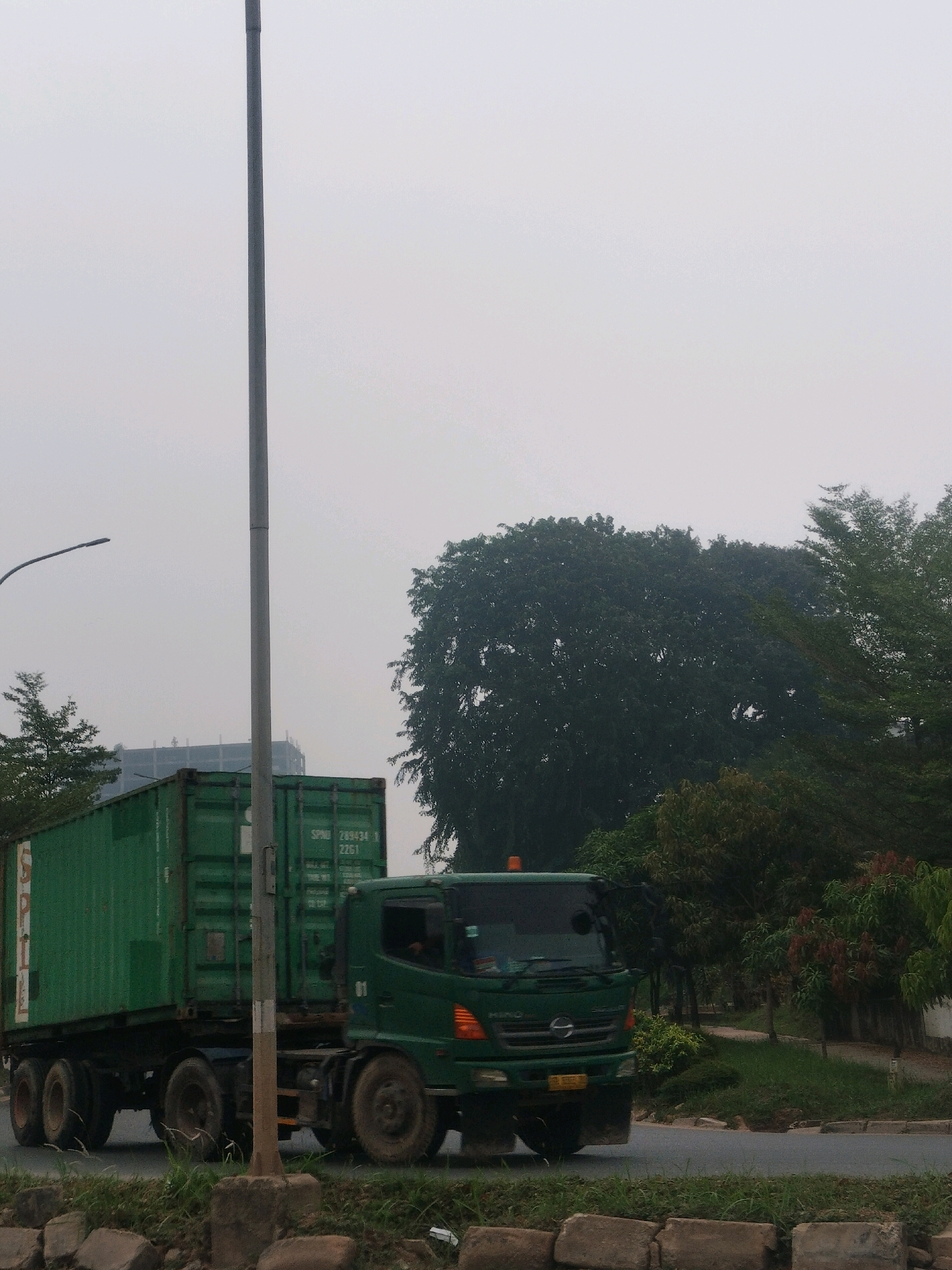 A green semi-trailer truck carrying a green shipping container travels along a paved road in Batam, Indonesia. The truck's cab is green, and it has orange turn signals. The shipping container has white lettering that appears to say "SPLL" and "289434 1 2261". In the background, there are several trees, some of which are tall and leafy. A street light pole stands to the left of the truck. The sky is overcast, suggesting a cloudy day. The road appears to be bordered by grass and some rocks.