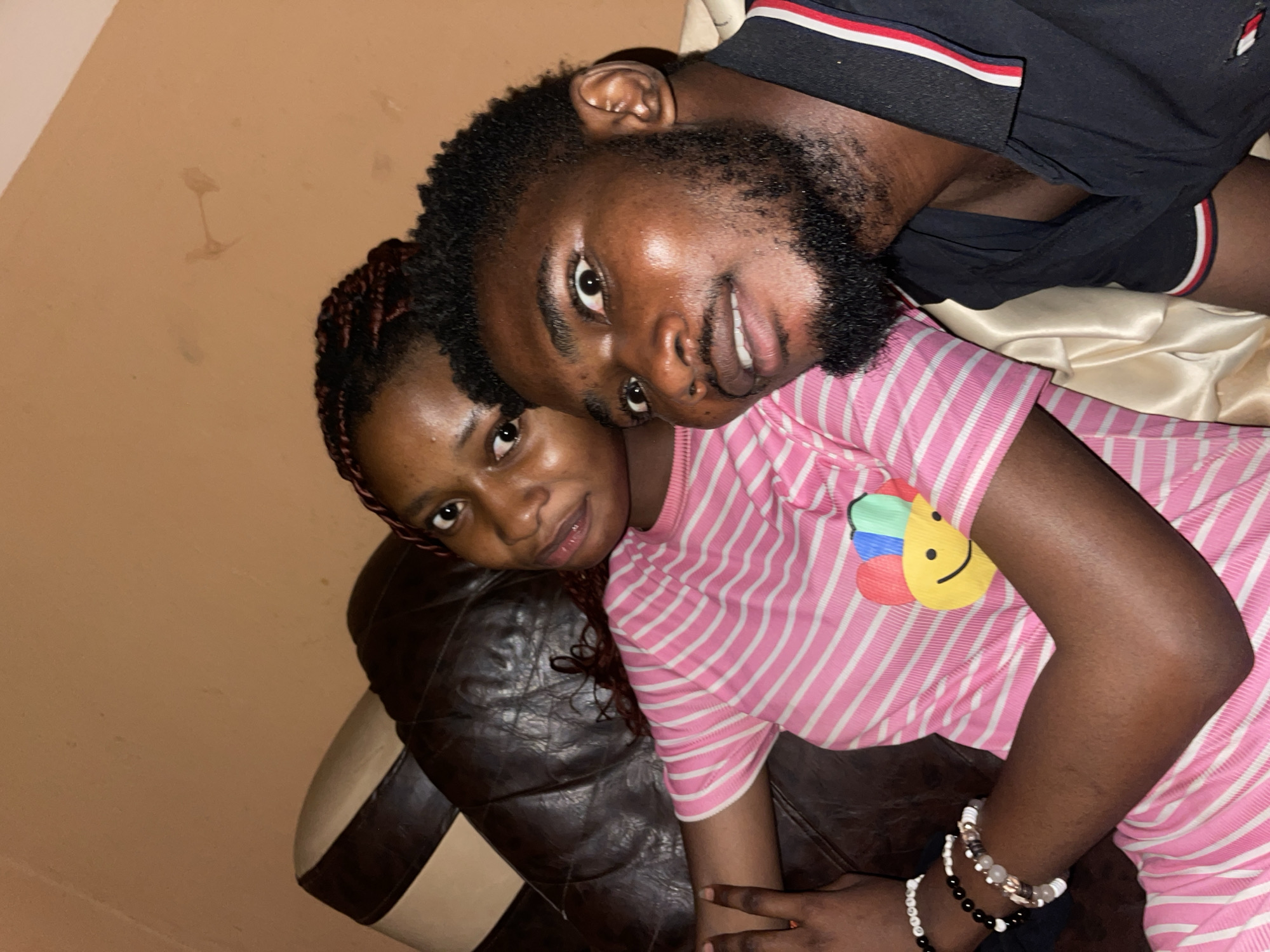 Two dark-skinned individuals are close together in what appears to be an indoor setting in Unguwar Gabas, Nigeria. The man has a beard and dark curly hair, while the woman has braided hair. The man is wearing a black shirt with a white and red stripe at the collar, while the woman is wearing a pink and white striped shirt with a smiley face decoration on it. She is also wearing a beaded bracelet. They are sitting on a dark brown leather surface. The walls are a light peach color.