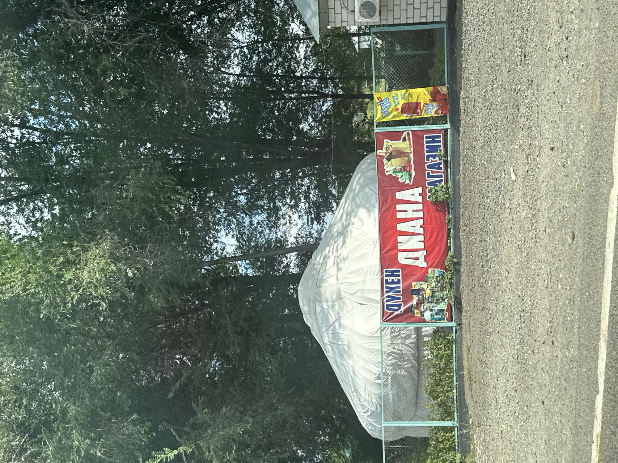 An outdoor scene in Көлбай, Kazakhstan features a large, white yurt covered in a tarp, situated behind a metal fence. In front of the yurt, a red banner displays Cyrillic text and images of bottles and products. To the right of the red banner, a yellow banner with Cyrillic text and an image of fruit is also visible. A brick building with an air conditioning unit is present on the right side of the image. Trees and foliage surround the area, and the ground is covered in gravel.