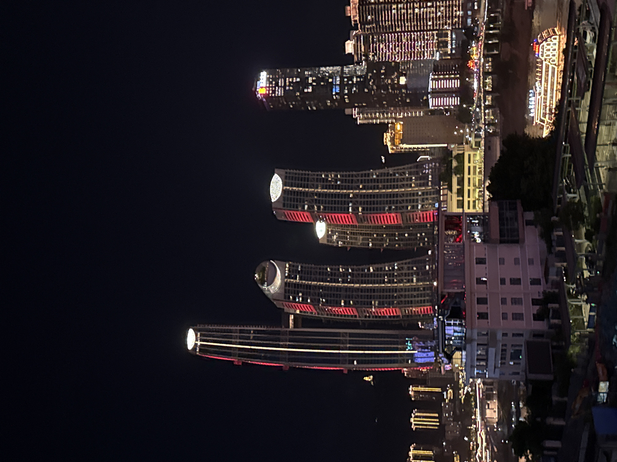 The image captures a nighttime view of a modern city skyline, likely in Yuzhong District, China, with several illuminated skyscrapers dominating the scene. The tallest skyscraper on the left is a slender, modern building with a distinctive illuminated top and vertical red and yellow lights running its length. To its right are two curved skyscrapers, also lit with red vertical lines. Further to the right, a more rectangular skyscraper is visible, punctuated by numerous lit windows and a red and blue illuminated sign at its peak. The background is filled with a dense collection of smaller illuminated buildings and structures, suggesting a bustling urban environment. In the foreground, a multi-story building with a flat roof and numerous windows is visible, with some areas lit from within. Below this building, trees and what appear to be covered parking areas with some vehicles can be seen. A large, brightly lit boat with multiple decks is navigating the water in the lower right portion of the image. The overall impression is a vibrant and dynamic urban landscape at night.
