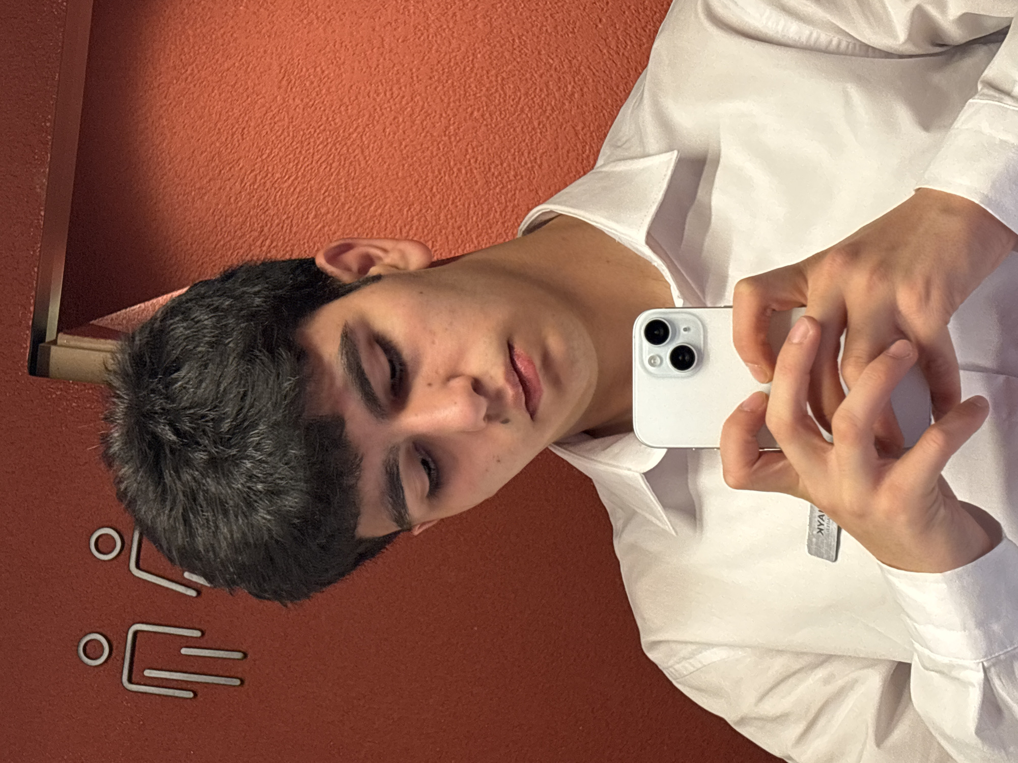 A young man in a white dress shirt is taking a selfie in a bathroom with red walls. The restroom has a simple icon of a person on the wall. It is not possible to confirm the location as Yerevan, Armenia based on the visual information.