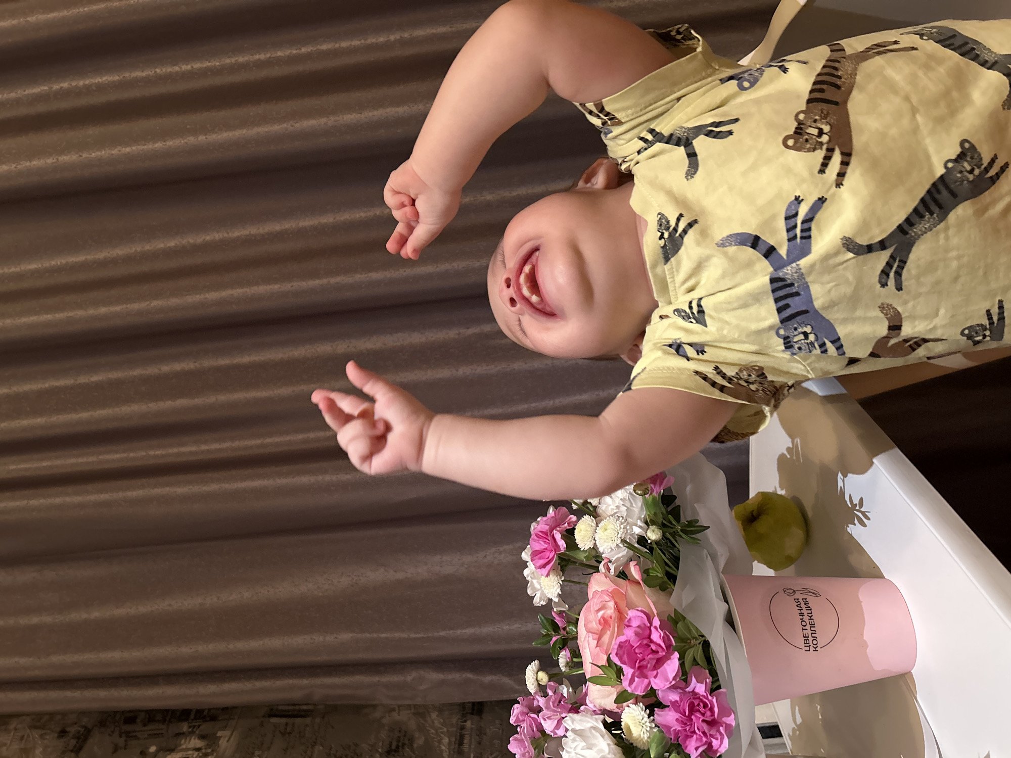 A baby in a yellow t-shirt with animal print is upside down, positioned above a bouquet of pink and white flowers and a green apple. The baby is smiling, with visible gums and teeth. The bouquet is in a pink cylindrical container labeled in Russian. A light-colored surface is behind the bouquet. In the background, there is a brown curtain. This image was likely taken in an indoor setting, potentially in the city of Chaikovsky, Russia.