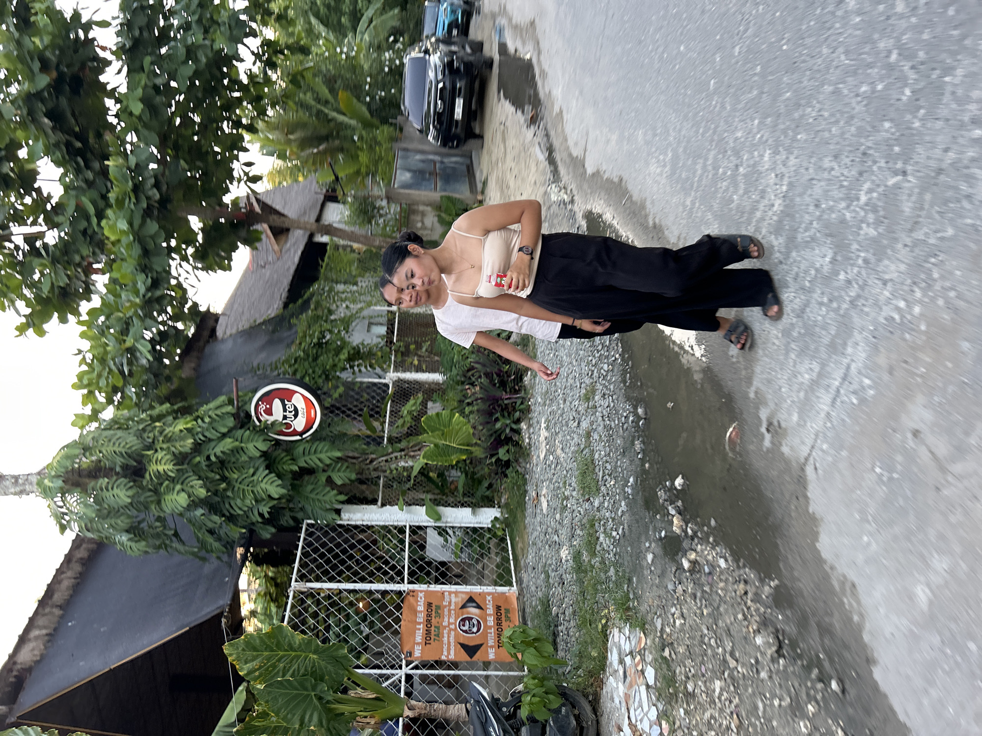 An outdoor, slightly tilted shot captures two young women standing on a road in General Luna, Philippines, likely during late afternoon or early evening. The foreground features a woman wearing a beige camisole, black wide-leg pants, and dark sandals, looking towards the camera with a relaxed expression. She wears a watch and holds a small red and white packet in her hand. Partially obscured behind her to the left is another person in a white t-shirt.
To their left, a metal fence separates the road from a lush, green area featuring various tropical plants and large-leafed vegetation. Behind the foliage, a building with a dark, possibly thatched roof is visible, suggesting a cafe or local establishment. A black SUV is parked further down the road in the background.
Visible details include a circular sign with a white and red logo, displaying the text "Outer." An orange rectangular sign attached to the fence reads: "WE WILL BE BACK TOMORROW 7AM - 3PM," followed by a menu listing "Pancakes, Bacon, Eggs & Rice bowl SM." Below this, another similar section of the sign repeats: "WE WILL BE BACK TOMORROW 3PM." The ground on the right consists of a grey concrete path with some puddles, bordering a gravel and dirt area. A motorcycle is partially visible near the bottom left corner by the fence.