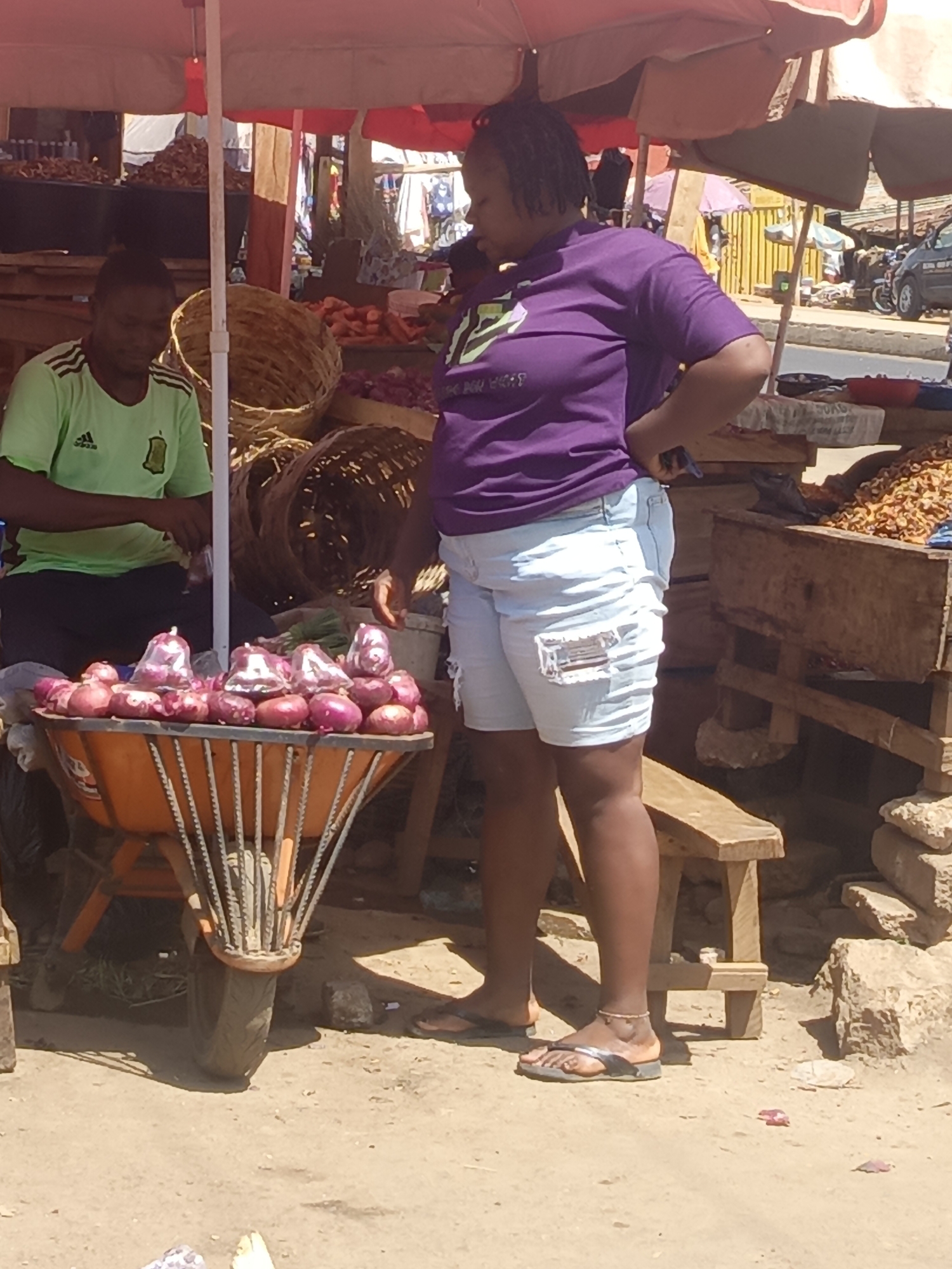 In Akwanga, Nigeria, a street market scene unfolds under the shade of a large, red-trimmed umbrella. A man wearing a green soccer jersey and black pants sits behind a display of woven baskets, packaging items. A woman in a purple shirt and light blue denim shorts stands next to a wheelbarrow filled with red onions, some bagged, seemingly engaging with the man. The wheelbarrow is orange and its metal frame is visible. A wooden bench sits nearby. In the background, other stalls and vendors are visible, along with parked cars, indicating a bustling market environment.
