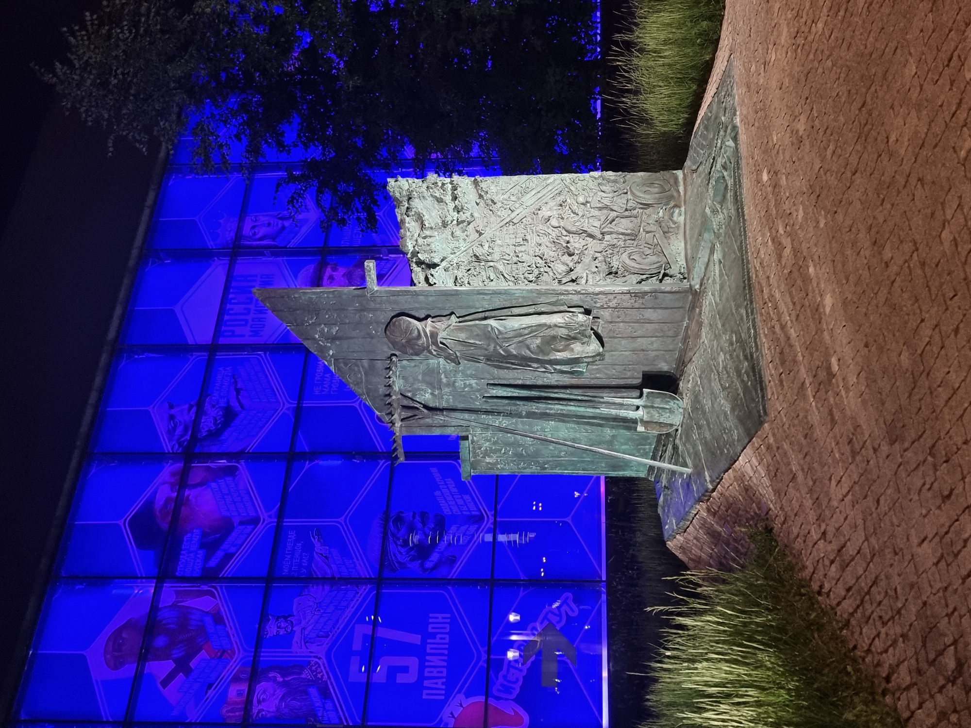 The image captures an outdoor scene at night, illuminated by intense blue lighting emanating from a building facade. In the foreground, a bronze sculpture stands on a paved area, made of red bricks laid in a circular pattern. The sculpture depicts agricultural tools, a shovel and a rake, leaning against a textured, wall-like structure. A cloak hangs from this structure. To the right of the sculpture, tall grass grows, and a dark tree looms against the blue-lit building. The building's facade is covered in a grid of illuminated panels, each displaying images and text in a hexagonal format. Some panels show portraits of historical figures, while others feature text such as "РОССИЯ МОЯ ИСТОРИЯ" (Russia My History) and the number "57" above "ПАВИЛЬОН" (Pavilion). The overall impression is a dramatic and possibly commemorative display, likely at an exhibition or memorial site. The specific location is likely the VDNKh (Exhibition of Achievements of National Economy) in Moscow, Russia, given the architecture and the content of the illuminated panels which often feature historical and national themes.