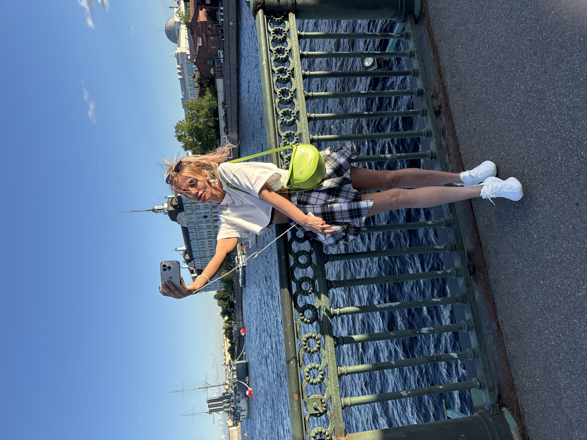 A young Caucasian woman with blonde hair pulled back in a ponytail, wearing sunglasses perched on her head, is taking a selfie on a bridge in Saint Petersburg, Russia. She is wearing a white crop top, a black and white plaid mini skirt, and white sneakers. A bright green shoulder bag is slung across her body. She is holding a silver smartphone in her right hand, with white earbuds in her ears. The background shows a wide river with a historical ship docked on the left side, and buildings with distinctive architecture along the embankment. The sky is a clear blue, suggesting a sunny day. The bridge has a dark green, ornate metal railing.